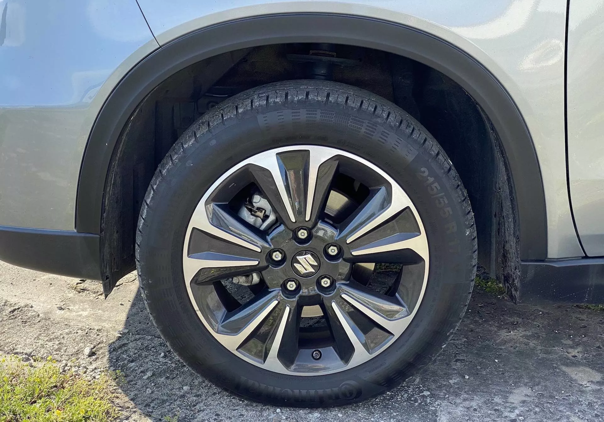 Zblízka pohľad na koleso Suzuki Vitara 2018, ktoré zobrazuje koleso zliatinovým dizajnom s viacerými kovovými a tmavšie natretými lúčmi. Značkový stredový kryt zakrýva matice kolies a cestná pneumatika obsahuje detailný vzor behúňa a bočné označenia. Pozadie obsahuje časť blatníka a bočného panelu vozidla v neutrálnej, lesklej farbe, snímaný z mierne naklonenej perspektívy od úrovne zeme.