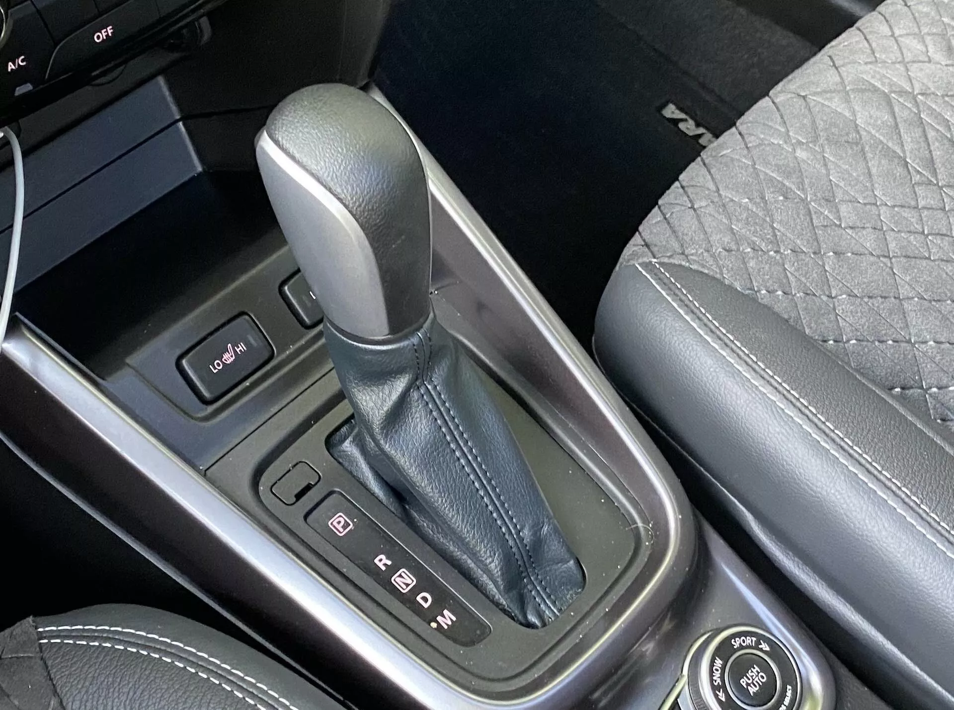 Detailný pohľad na stredovú konzolu Suzuki Vitara 2018, zameraný na páku radenia v polohe 'P' pre parkovanie, obklopenú kovovým krytom s jasnými ukazovateľmi. Naľavo je viditeľný ovládací panel s dvoma tlačidlami a okraj obrazovky. Stredová konzola je vyrobená z vysoko kvalitných materiálov a na pravej strane je časť predného sedadla s prešívaným dizajnom. K dispozícii je aj otočný ovládač pre infotainment funkcie, čo poukazuje na pokročilé technológie a luxusné prvky vozidla.