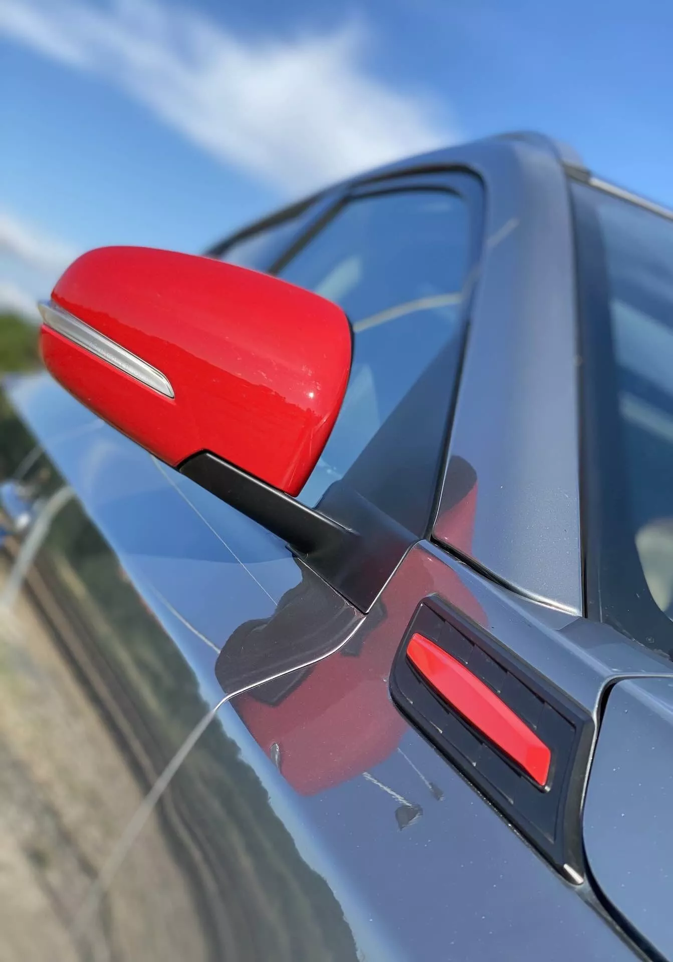 Zblízka pohľad na spätné zrkadlo na strane vodiča Suzuki Vitara 2018, ktoré je zvýraznené červenou farbou voči tmavej farbe karosérie auta. Zrkadlo je pripevnené soštíhleným čiernym držiakom, čo naznačuje aerodynamickú účinnosť. Záber zachytáva zrkadlo zo šikmého uhla mierne spredu, z pohľadu mierne nadol, s červenou lištou na dverách. Pozadie je rozmazané, čím sa zdôrazňujú detaily zrkadla a dizajnu dverí auta.