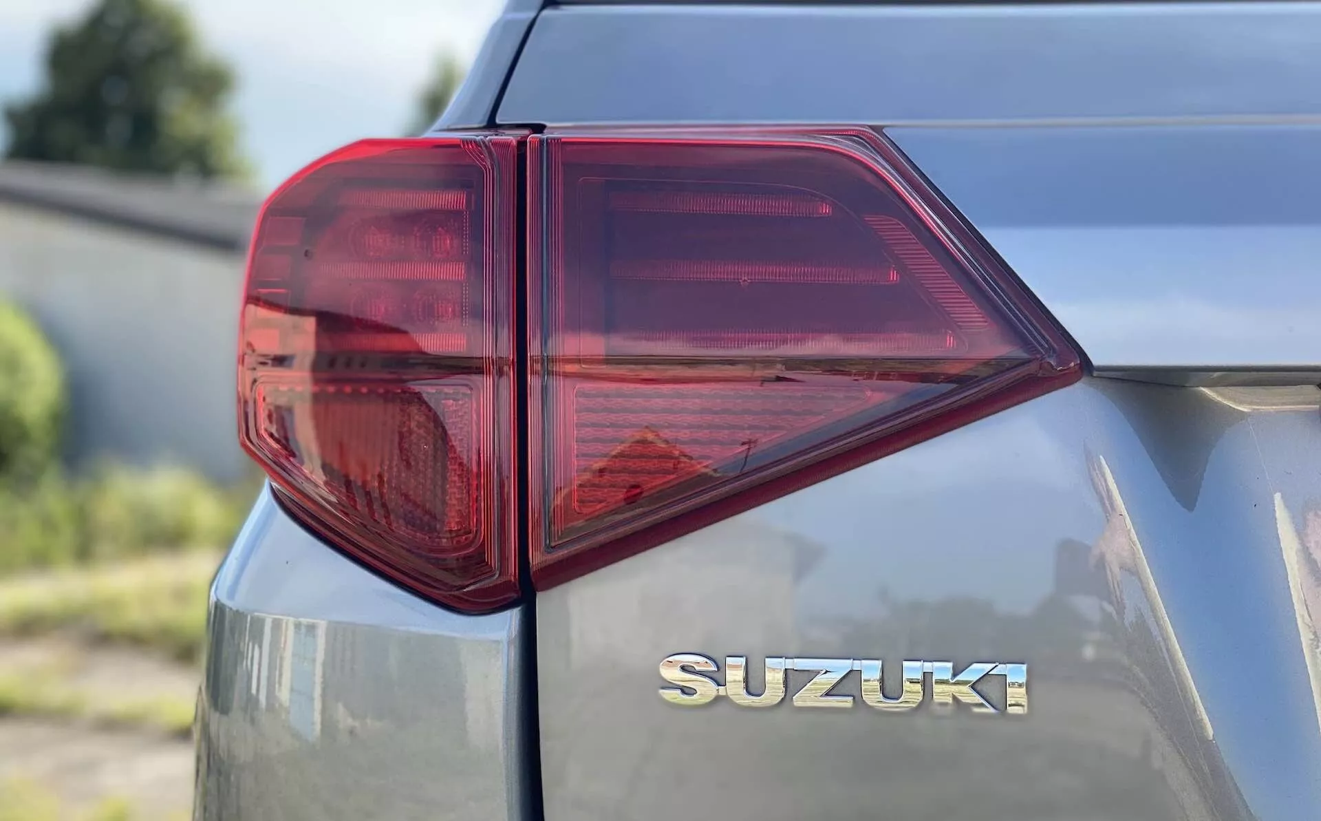 Detailný pohľad na zadnú ľavú stranu modelu Suzuki Vitara 2018 so zameraním na zadné svetlo. Perspektíva z troch štvrtín zboku, urobená z nízkej až strednej výšky, zobrazuje charakteristicky tvarované LED zadné svetlo s ostrými uhlami a viacerými komorami pre funkcie ako brzdové svetlo, smerovka a prípadne hmlové svetlo. Prevažne červené zadné svetlo s jasnou sekciou pravdepodobne pre spiatočku, zdôrazňuje modernú automobilovú osvetľovaciu technológiu. Zadné veko obsahuje chrómový znak 'SUZUKI' umiestnený pod zadným sklom, čo zdôrazňuje čisté a hladké značenie. Kovovo lakovaný povrch odráža svetlo, čo naznačuje dobre udržiavaný alebo novší povrch vozidla.