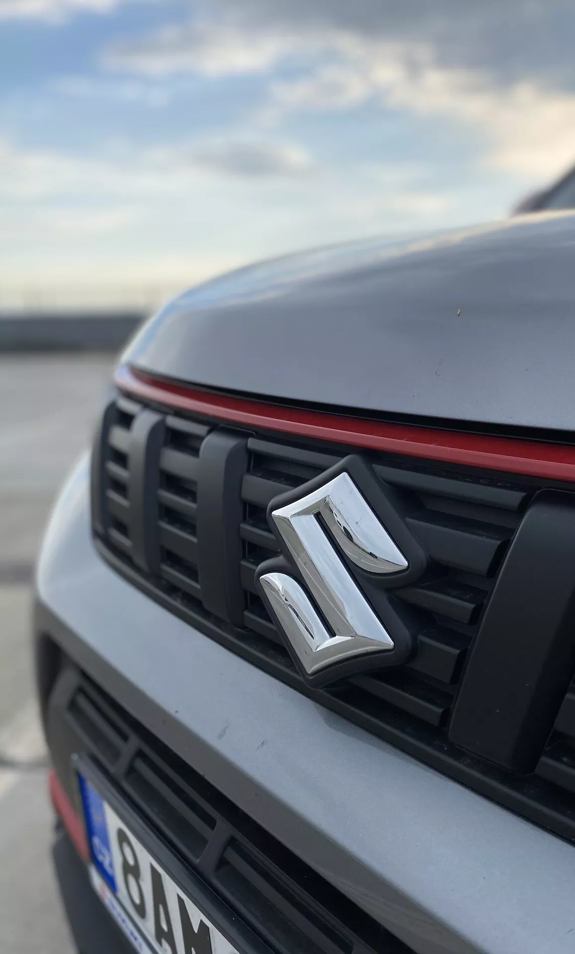 Detailný pohľad z nízkeho uhla na prednú časť vozidla Suzuki Vitara 2018. Obrázok výrazne zobrazuje emblém výrobcu umiestnený v strede mriežky, ktorá má horizontálne lamely a červený lem cez hornú hranu, čo naznačuje športovejší dizajn. Kapota a časti auta sú rozmazané, čo zdôrazňuje biely alebo svetlý povrch s rozpoznateľným prehybom. Pozadie je tiež rozmazané, čo naznačuje vonkajšie prostredie, pravdepodobne na parkovisku, v ranných alebo neskorých popoludňajších hodinách.