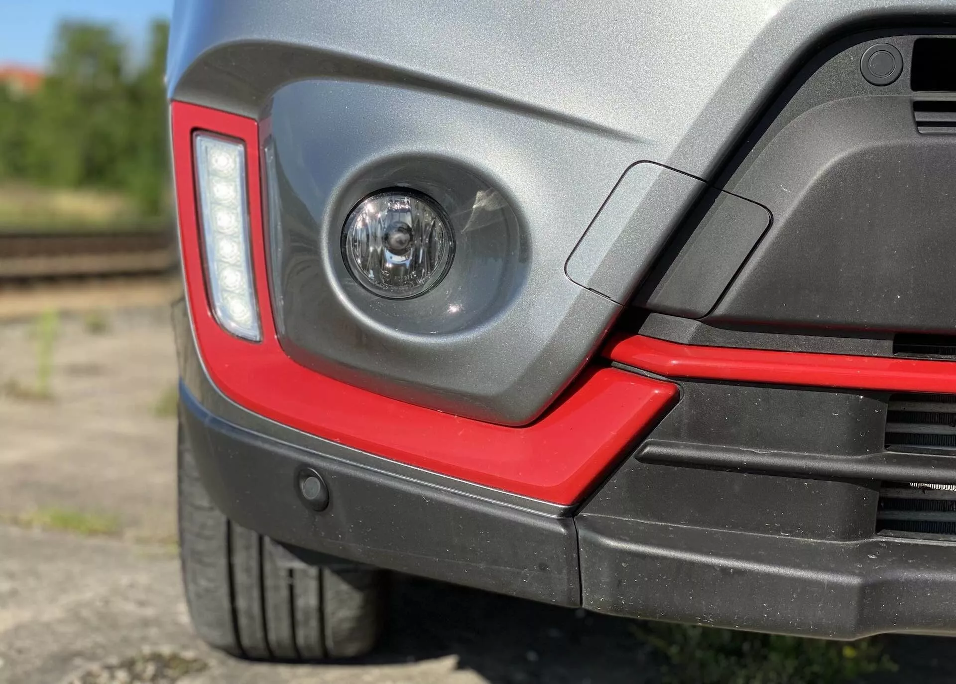 Detailný pohľad na zadný roh Suzuki Vitara 2018, ktorý zahŕňa čiré mlhové svetlo v zadnom nárazníku, moderný hranatý dizajn zadných svetiel, dvojfarebný zadný nárazník so športovým červeným akcentom, potenciálne parkovacie senzory, nízkoprofilovú pneumatiku s ochranným plastovým oblúkom, lesklý lak a rozmazané polopriemyselné pozadie s železničnými koľajami.