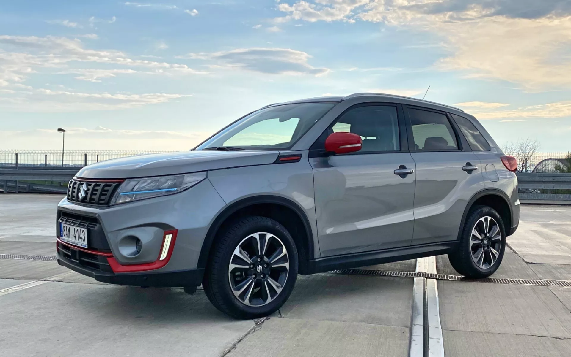 Obrázok zobrazuje Suzuki Vitara 2018, moderné kompaktné SUV, zaparkované vonku za denného svetla. Je zachytené z trojštvrťového predného pohľadu, zvýrazňujúceho predný aj bočný profil. Uhol je mierne nízky, na úrovni kľučiek dverí, čo naznačuje, že fotograf kľačal alebo sa ohýbal. Viditeľné detaily zahŕňajú prednú mriežku, svetlá, púzdro hmlových svetiel, emblém, bočné zrkadlá, zliatinové kolesá so zabudovanými smerovými svetlami, kľučky dverí, okenné lišty, líniu strechy a strešné lišty. Projektorové svetlá a elegantná anténa naznačujú pokročilé technológie. Moderné dizajnové prvky SUV naznačujú funkcie ako LED svetlá a možno aj systém bezkľúčového vstupu.