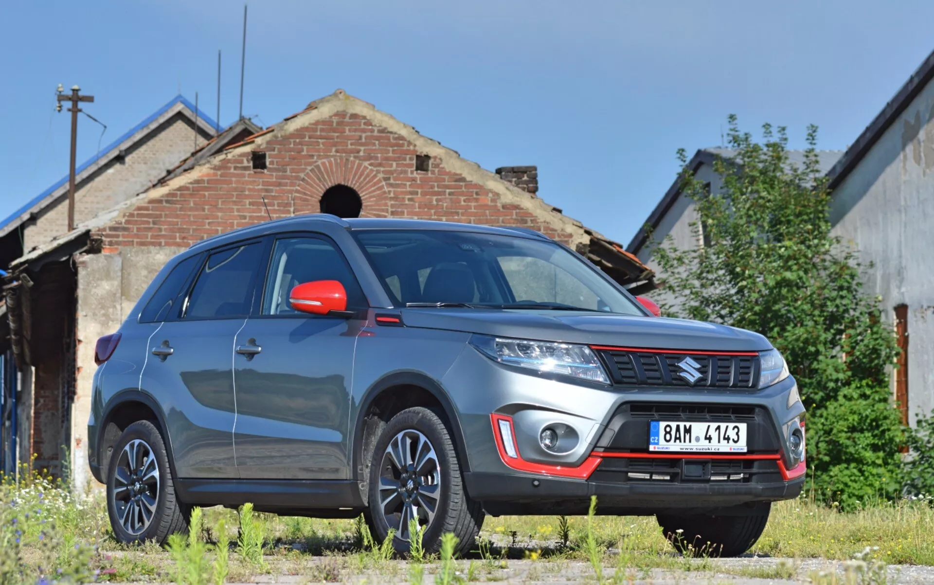 Obrázok zobrazuje SUV Suzuki Vitara 2018 z nízkej trojštvrťovej prednej perspektívy, zvýrazňujúcej pravú prednú stranu vozidla s výraznou svetlou výškou. Viditeľné časti zahŕňajú robustný predný nárazník s hmlovými svetlami a strieborným krytom, emblém značky na horizontálnej mriežke, výrazné línie kapoty, moderné svetlomety s možnou LED alebo HID technológiou, viaclúčové hliníkové disky, červené spätné zrkadlá s integrovanými smerovkami, svalnaté blatníky, reflexné čelné sklo a tónované okná. Vitara je zaparkovaná na nezpevnenom trávnatom povrchu pred starou budovou, čo pridáva industriálny alebo vidiecky podtext, naznačujúci vhodnosť pre vonkajšie prostredie.