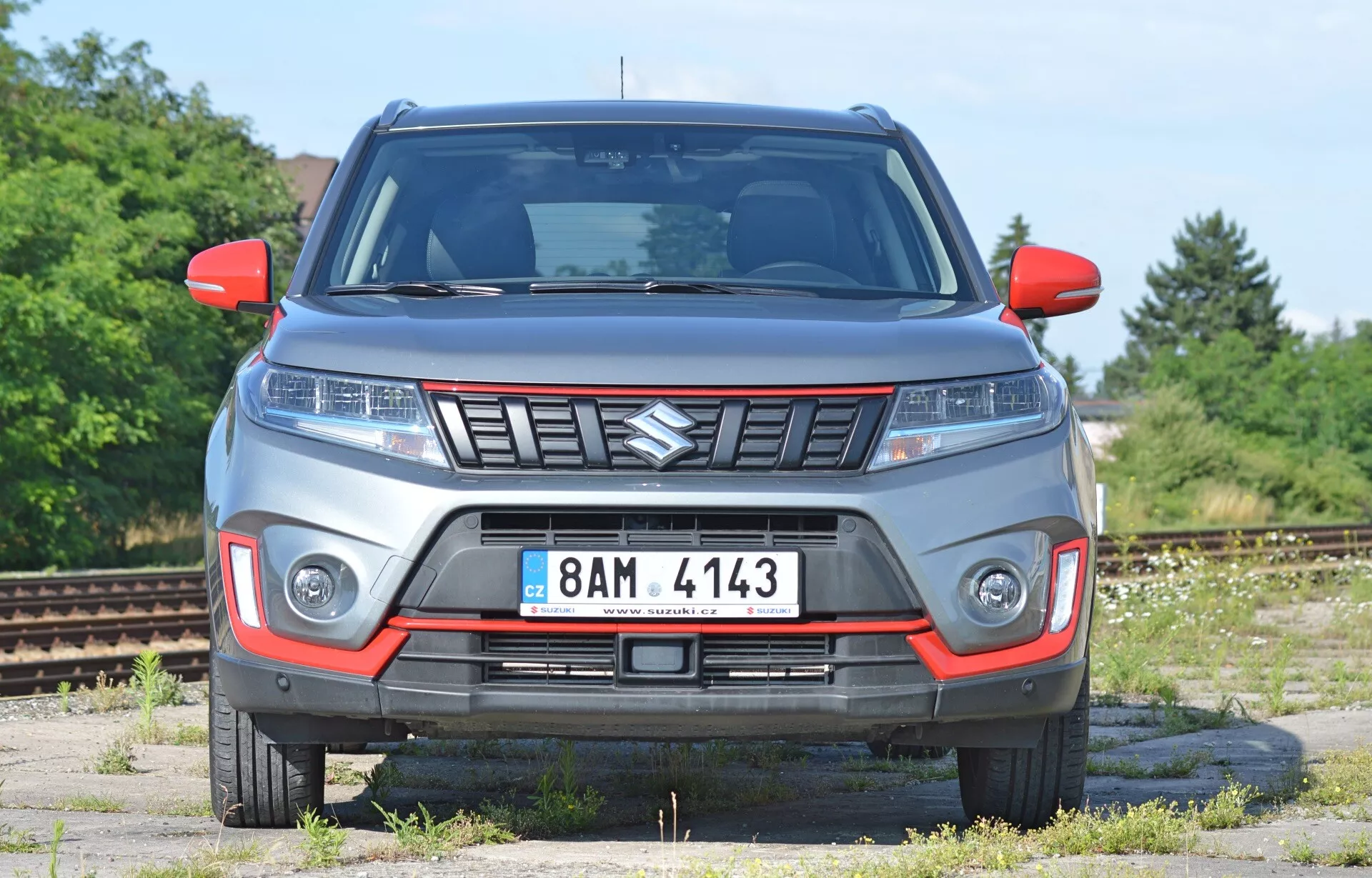 Čelný pohľad na Suzuki Vitara 2018 s mierne nízkym uhlom. Obrázok zvýrazňuje predné rysy vozidla, vrátane kapoty, mriežky (so znakom Suzuki), elegantných svetlometov (potenciálne LED) a osvetlenia na nárazníku. Vozidlo pôsobí robustne, čo naznačuje schopnosť jazdiť v teréne. Viditeľné sú smerovky v zrkadlách a predná poznávacia značka. Sivá karoséria s červenými akcentmi môže znamenať špeciálne vybavenie. Pozadie sa javí ako opustená priemyselná oblasť alebo železničný dvor, čo poskytuje jasný pohľad na dizajn vozidla.