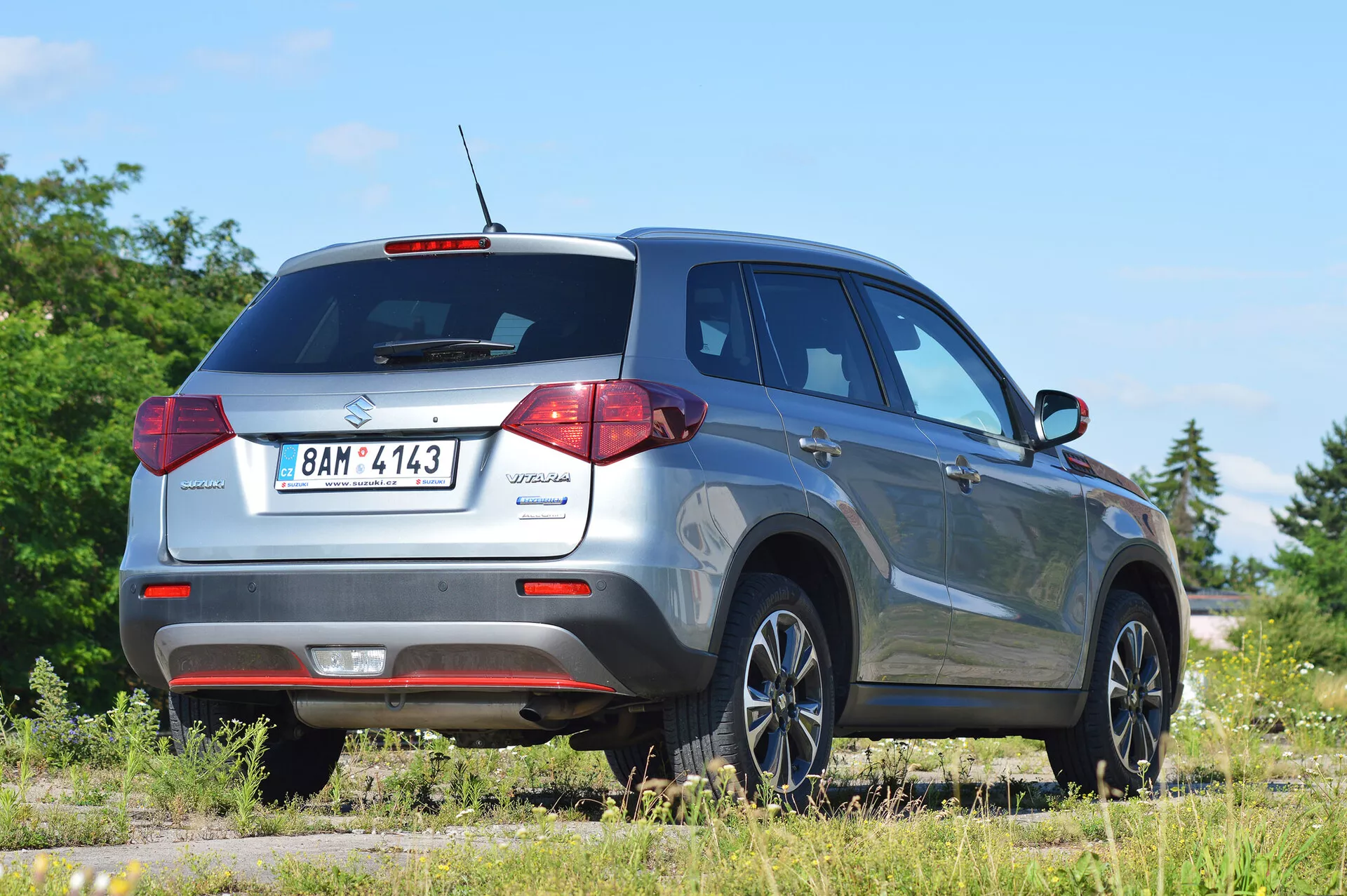 Suzuki Vitara 2018 zachytená z pravého zadného trojštvrťového uhla, zdôrazňujúci dizajn zadných svetiel, zadné okno a profil na strane spolujazdca. Obrázok zobrazuje moderné vybavenie kompaktného SUV, ako je strešná anténa pre technologické funkcie, zadné parkovacie senzory a štýlové zliatinové kolesá. Vozidlo je umiestnené na nepevnom povrchu v vidieckom prostredí, čo naznačuje jeho schopnosť off-road. Ideálne pre kategórie automobilov, SUV a obrázky tvoriace terénne vozidlá.