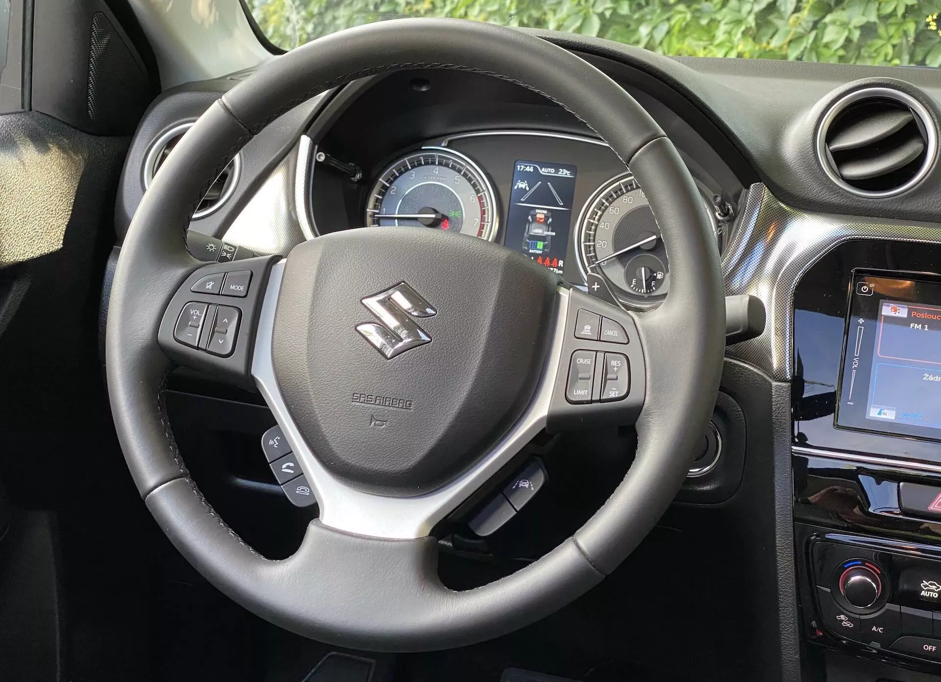 Interiérový obraz Suzuki Vitara 2018, zobrazujúci perspektívu vodiča so zameraním na volant a prístrojovú dosku. Volant, situovaný v strede obrazu, má opierky na palce a ovládače pre audio, hlasové povely a tempomat. Za volantom je prístrojový panel so zobrazeným analógovým rýchlomerom a čiastočne digitálnym displejom poskytujúcim informácie pre vodiča. Na pravej strane volante časť stredovej konzoly ukazuje dotykový infotainment systém, čo zdôrazňuje moderný vzhľad vozidla.