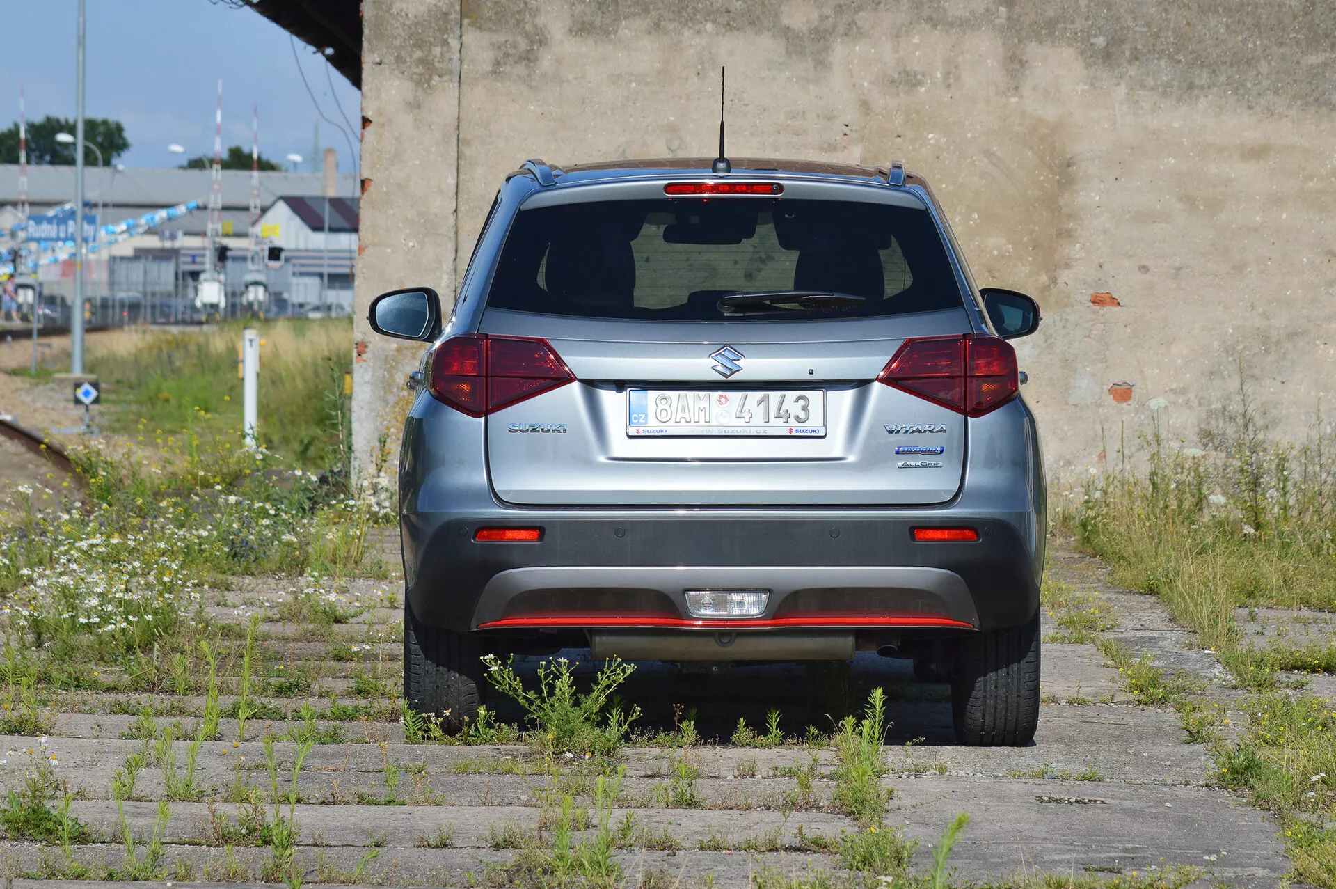Zadný uhol strieborného kompaktného SUV Suzuki Vitara 2018 zaparkovaného na trávnatej a betónovej ploche. Viditeľná je celá zadná časť, s centrálnou registračnou značkou, zadným stieračom, tretím brzdovým svetlom, štandardnými halogénovými koncovými svetlami, odrazkami na nárazníku, parkovacími senzormi a výfukovou rúrou na ľavej strane. Vozidlo sa nachádza v polozapustenom priestore s prerastenou vegetáciou a opotrebovanou stenou.