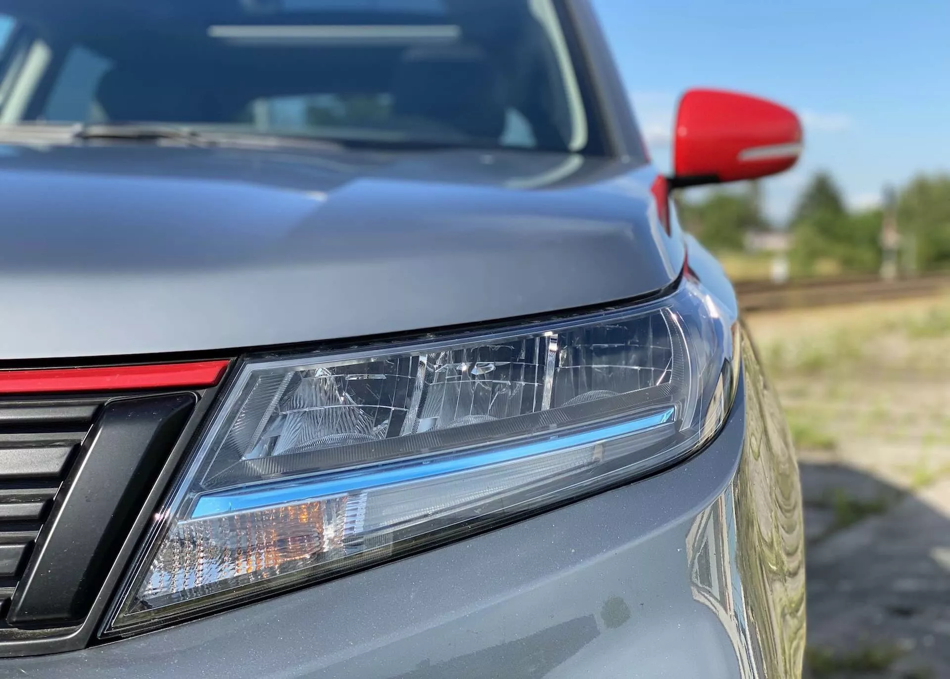 Detailný záber na predný ľavý roh vozidla Suzuki Vitara 2018, zameraný na detaily ako LED svetlomety s dennými svetlami, čiastočne viditeľná mriežka s vertikálnymi lištami, jemne sa zvažujúcu kapotu, kovový lak, kontrastné červené bočné zrkadlo pravdepodobne s integrovanými smerovkami a rozmazané pozadie, ktoré zdôrazňuje elegantný dizajn a aerodynamickú účinnosť.