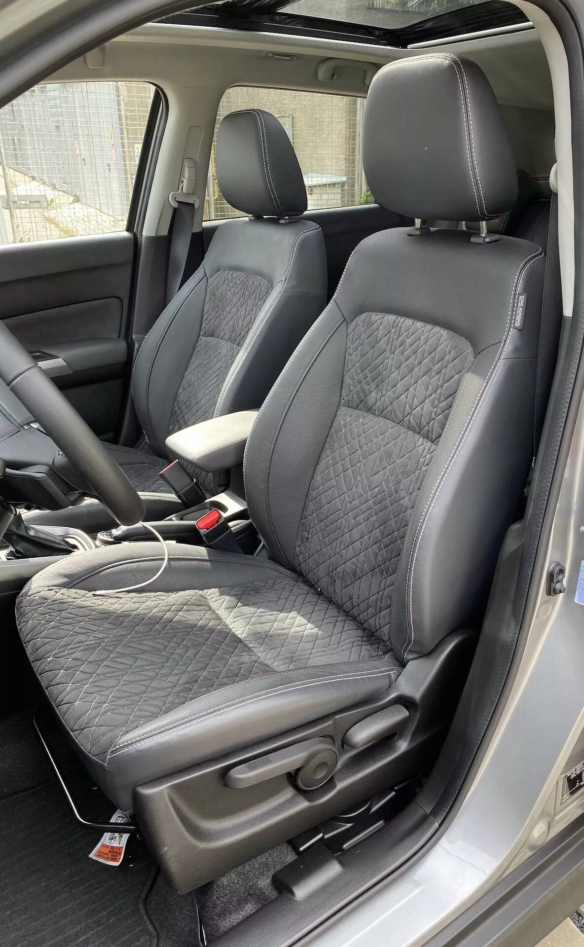 Interiér Suzuki Vitara 2018, zvýrazňujúci moderné predné sedadlá v štýle vedra z perspektívy mierne nad úrovňou sedadiel. Sedadlá sú čalúnené čiernou kožou s prešívaným diamantovým vzorom a hladkými koženými bokmi, ponúkajúc rovnováhu medzi pohodlím a športovosťou. Červený štítok na sedadlách naznačuje ďalšie funkcie, ako sú bočné airbagy alebo vyhrievané sedadlá. Viditeľná je aj stredová konzola s koženou opierkou ruky, ako aj nabíjací kábel na sedadle spolujazdca, čo naznačuje prítomnosť nabíjacích portov. Zapadania bezpečnostných pásov sa zhodujú s čiernym interiérom. Viditeľná je aj časť prístrojovej dosky a stĺp B, čo naznačuje priestrannú kabínu.