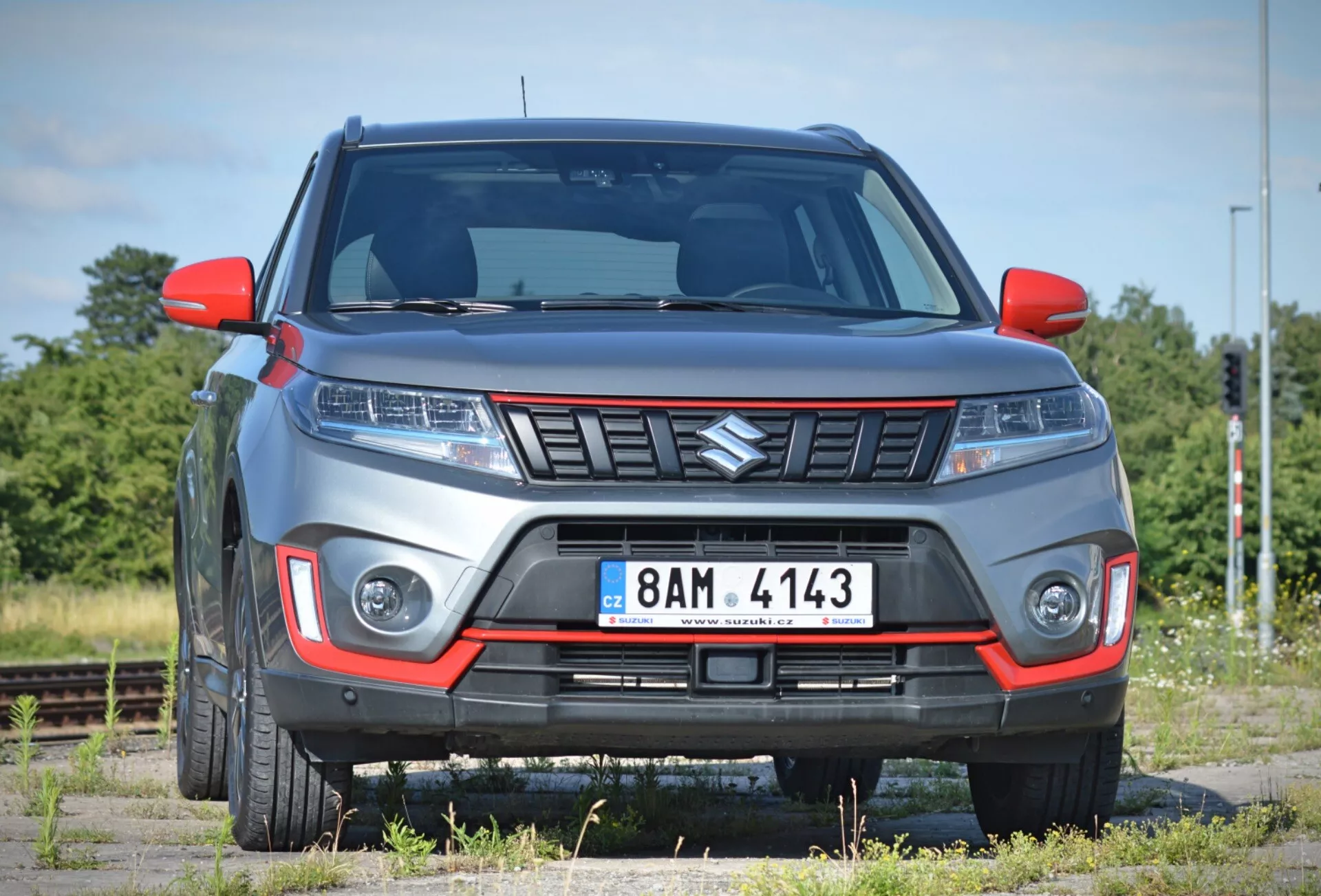 Obrázok ukazuje moderné 2018 Suzuki Vitara SUV pod miernym predným uhlom, zvýrazňujúc prednú časť a časť strany spolujazdca na pozadí riedkeho krajinného prostredia s železničnými traťami. Auto, zaparkované na povrchu vyzerajúcom ako štrkový, má výraznú mriežku s uhlovým dizajnom, elegantné svetlomety pravdepodobne s technológiou LED alebo HID a integrované denné svetlomety alebo hmlové svetlá v prednom nárazníku. Nárazník obsahuje kontrastnú vložku naznačujúcu terénnu alebo športovú úroveň výbavy, a viditeľná svetlá výška posilňuje jeho charakter SUV. Zvýšené línie na kapote prispievajú k agresívnemu štýlu, zatiaľ čo bočné zrkadlá a blatníky ukazujú moderný dizajn. Čiastočne viditeľné zliatinové kolesá dopĺňajú robustný vzhľad. Fotografia, urobená z blízkej vzdialenosti, umožňuje podrobné preskúmanie týchto charakteristík.