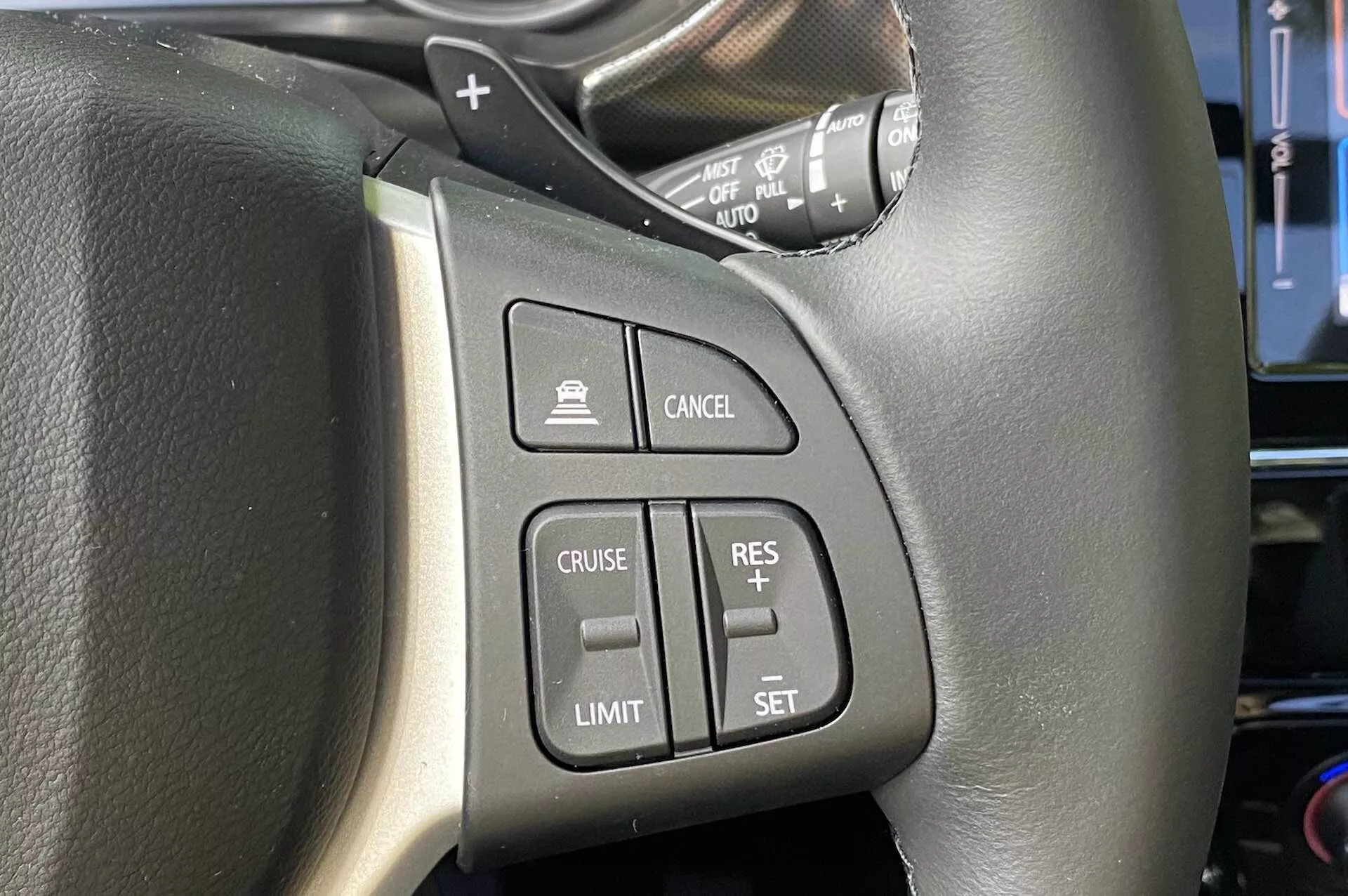 Detailný pohľad na ovládacie prvky na volante Suzuki Vitara 2018, zameraný na ľavú stranu. Čierny kožený volant s viditeľným šitím má ľahko dostupné tlačidlá vrátane aktivácie tempomatu, aktivácie obmedzovača rýchlosti, nastavenia/upravenia tempomatu a tlačidla na zrušenie. Na obrázku je tiež vidieť ovládaciu páčku pravdepodobne pre smerové svetlá a svetlomety, pohľad z pozície vodiča.