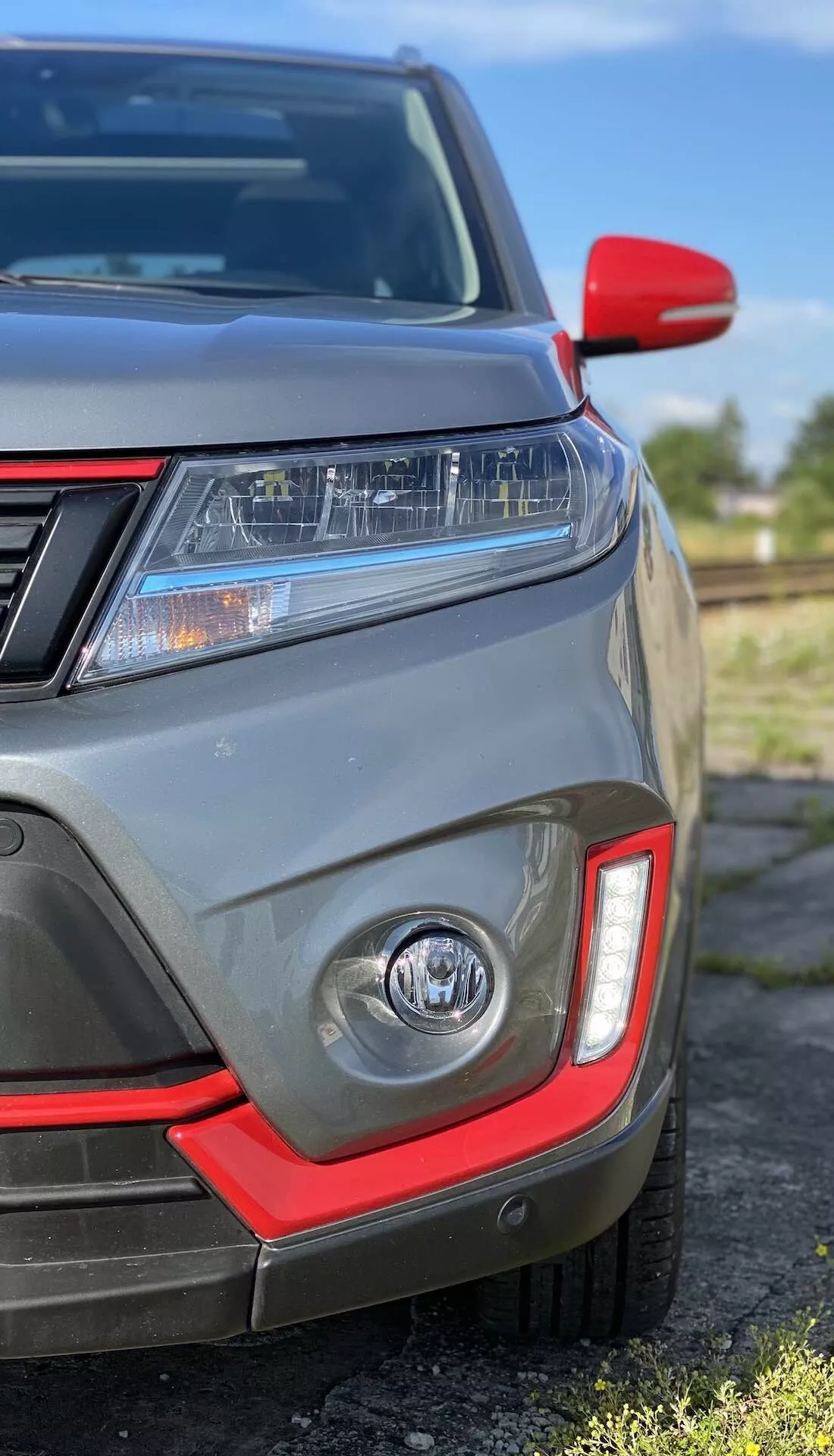 Detailný záber na predný ľavý kvadrant Suzuki Vitara 2018 zobrazujúci LED svetlomety, denné svietenie, hmlovku s červeným akcentom, parkovacie senzory, čiastočnú prednú mriežku, červeným krytom spätného zrkadla a metalickej šedej farby.