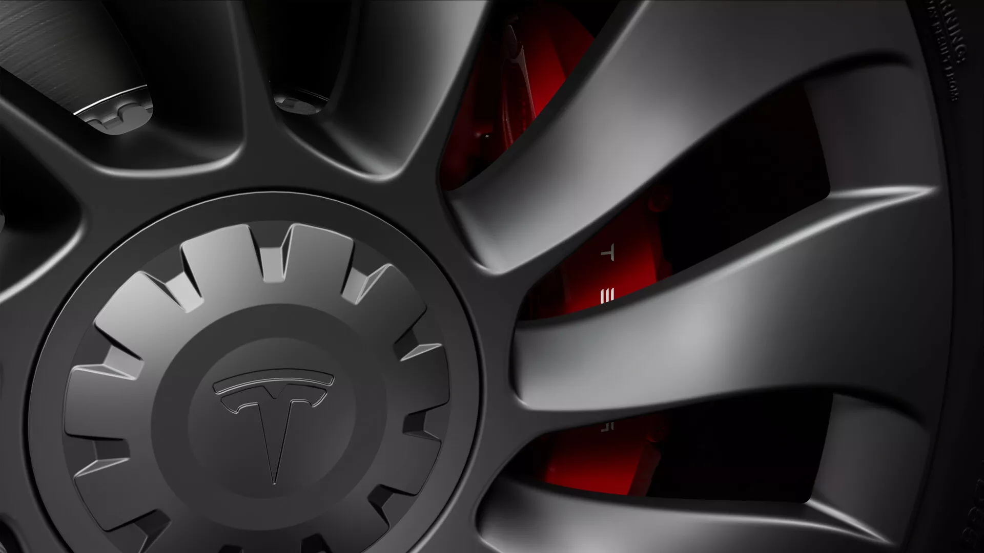 Detailný pohľad na koleso Tesla Model-3 2019 (1. Generácia), ktoré ukazuje vysokovýkonný viacramenný ráfik s tmavým matným povrchom, naznačujúci športový alebo prémiový vzhľad. Zložitý dizajn turbíny pomáha pri chladení bŕzd a zlepšuje aerodynamiku. Stredový kryt je zámerne zakrytý, aby sa ukryla značka. Za lúčmi je čiastočne viditeľný červený brzdový strmeň, signalizujúci vysokovýkonný brzdový systém. Chladený brzdový kotúč, navrhnutý na efektívne rozptyľovanie tepla, je čiastočne viditeľný, a okraj nízkoprofilovej pneumatiky je sotva viditeľný. Dramatické osvetlenie zvýrazňuje textúru a tvar, čím zdôrazňuje technologickú premyslenosť komponentov kolesa a bŕzd.