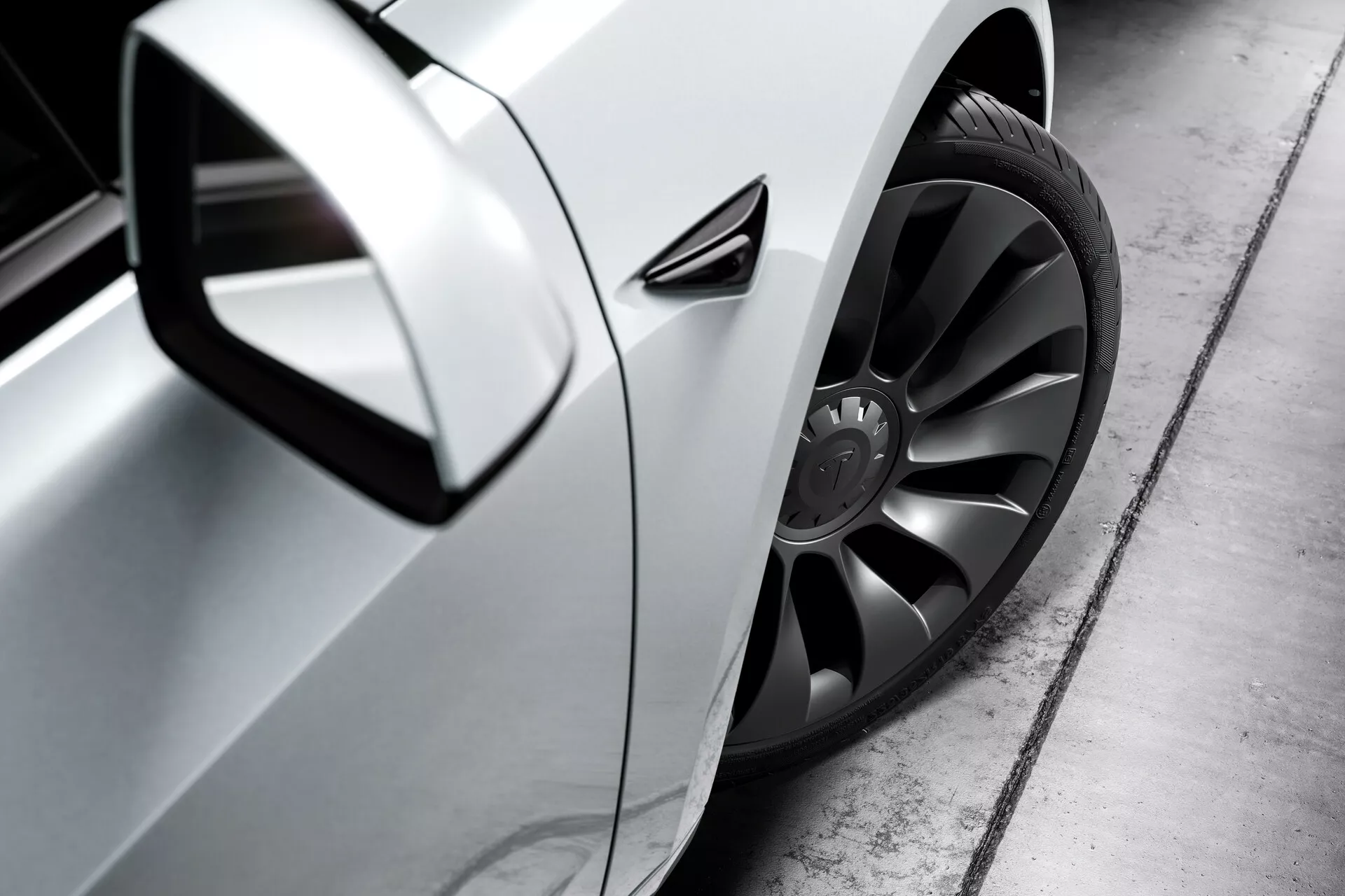 Detailný bočný pohľad na strieborný Tesla Model 3 (2019, 1. generácia) zameraný na predné koleso, oblasť dverí vodiča a bočné zrkadlo. Fotografia, urobená z nízkeho uhla blízko úrovne zeme, vyzdvihuje veľké, štýlové koleso z ľahkých zliatin s tmavým viacpaprskovým dizajnom a pneumatikou s nízkym profilom, elegantný aerodynamický bočný panel s kovovým povrchom a aerodynamicky integrované bočné zrkadlo. Kľučka na dverách sa zdá byť v línii s karosériou, čo naznačuje pokročilú aerodynamiku, zatiaľ čo malý ventil za predným kolesom môže slúžiť na chladenie bŕzd, aerodynamiku alebo estetiku. Dobre definovaný odraz na paneli naznačuje vysokú kvalitu laku.