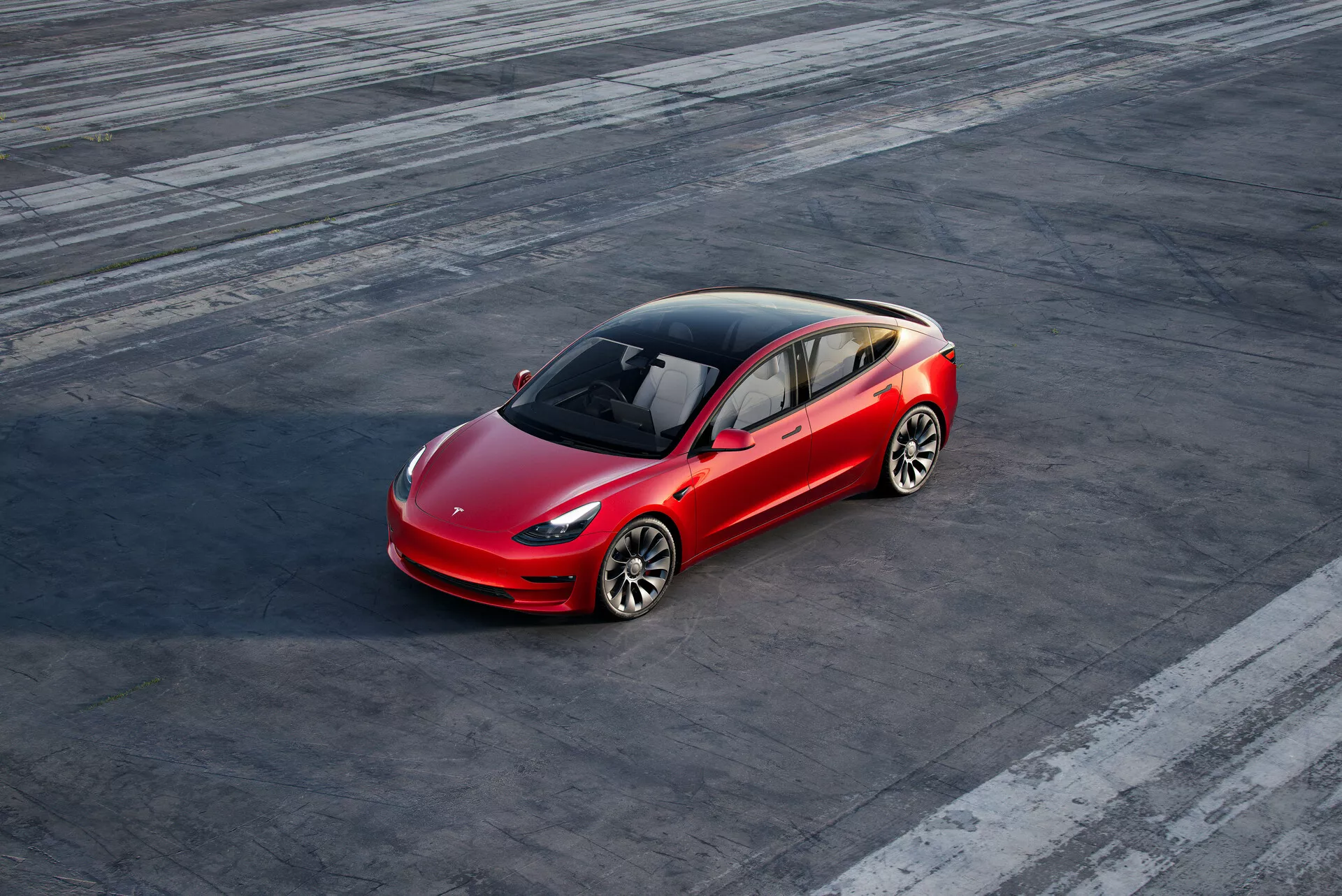 Snímka z vysokého uhla červeného štvordverového sedanu Tesla Model 3 (2019, 1. generácia), zaparkovaného na textúrovanej asfaltovej ploche s viditeľnými stopami pneumatík, čo naznačuje predchádzajúce jazdné manévre. Auto je umiestnené mierne doprava od stredu rámu a viditeľné z perspektívy prednej strany z troch štvrtín, s výhľadom na vodičovu stranu a čiastočný zadný pohľad. Aerodynamický dizajn, s výraznou kontúrovou líniou, panoramatickou sklenenou strechou, viacsílkovými hliníkovými kolesami, LED svetlami a minimalistickou prednou mriežkou zdôrazňuje pokročilú technológiu a dizajn vozidla.