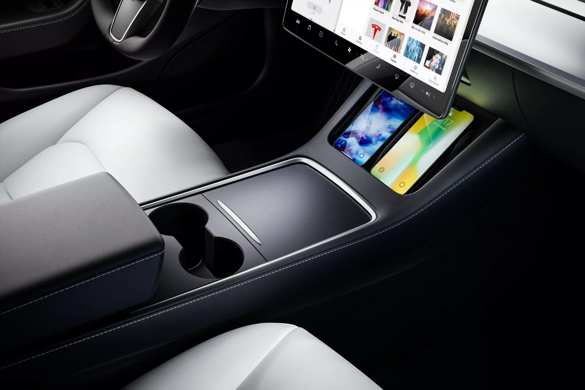 Záber zblízka na stredovú konzolu a predné sedadlá modelu Tesla Model 3 z roku 2019. Obraz, vytvorený z pohľadu zhora pod uhlom, zvýrazňuje elegantný, minimalistický dizajn stredovej konzoly s veľkým, horizontálne orientovaným dotykovým displejom. Infotainment systém zobrazuje multimediálne možnosti, ako je integrácia smartfónu a GPS navigácia. Pod dotykovým displejom sa nachádza rovný povrch, dva držiaky na poháre a otvorený úložný priestor. Tmavé tóny konzoly kontrastujú so svetlou kožou sedadiel, čo odráža luxusný a moderný dizajn. V ľavom dolnom rohu je viditeľný pohľad na ovládacie koliesko, pridávajúce k technologickému nádychu moderného interiéru auta.