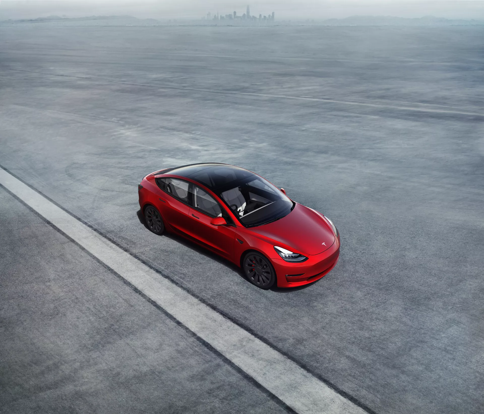 Letecký pohľad na odvážne červený Tesla Model 3 2019 (1. generácia) zaparkovaný na rovnom šedom povrchu. Elegantný štvordverový sedan má aerodynamický profil, šikmú strechu spájajúcu sa s kufrom a unikátne zliatinové kolesá. Zapustené kľučky dverí naznačujú aerodynamickú účinnosť. Vysoký stupeň tónovania okien, absencia viditeľného výfukového systému a moderné tenké svetlomety poukazujú na pokročilú technológiu a potenciálne elektrický alebo hybridný pohon. Auto je umiestnené paralelne s rovnou čiarou, čo naznačuje usporiadané umiestnenie.
