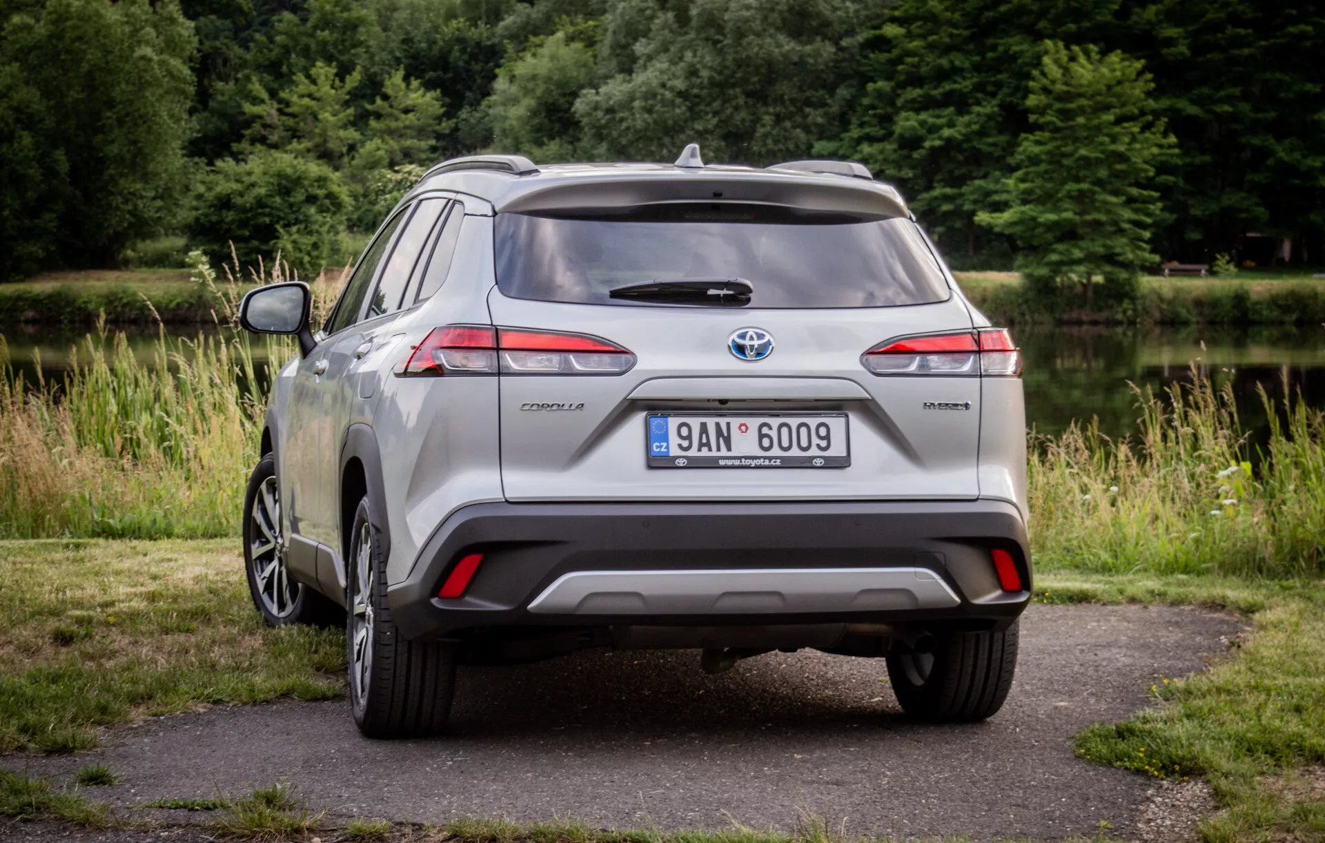 Strieborný Toyota Corolla Cross z roku 2022 zaparkovaný na dlažbe so zeleňou na pozadí, ktorý predstavuje jeho LED zadné svetlá, zadný spojler, zliatinové koleso s viacerými paprskami a označenie hybrid v prirodzenom dennom svetle