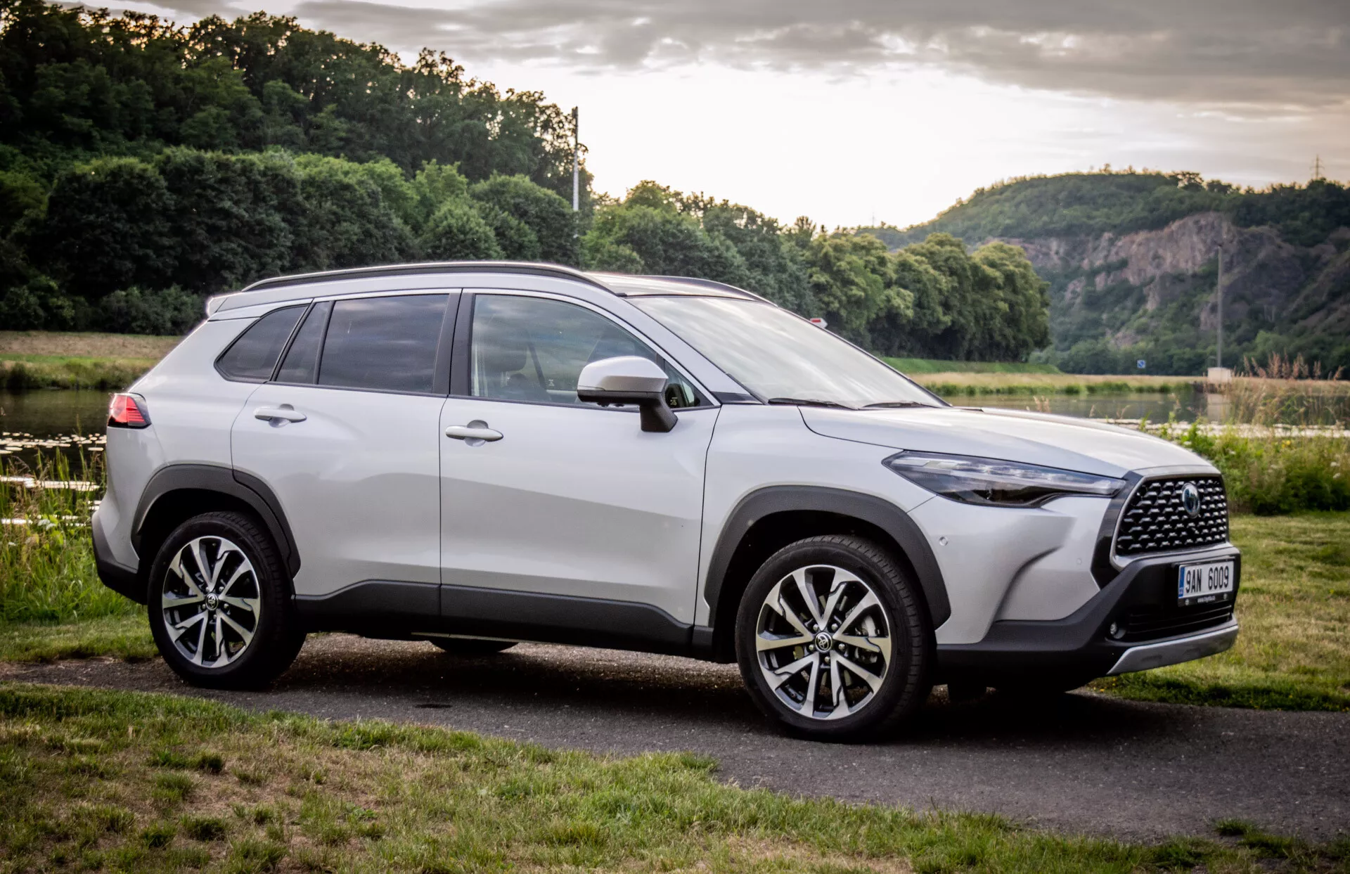 SUV Toyota Corolla Cross 2022 zaparkované vonku, predvádzajúce svoj moderný dizajn z čelno-bočného uhla so zameraním na mriežku chladiča, zliatinové kolesá a bočný profil na pozadí prírodnej scenérie