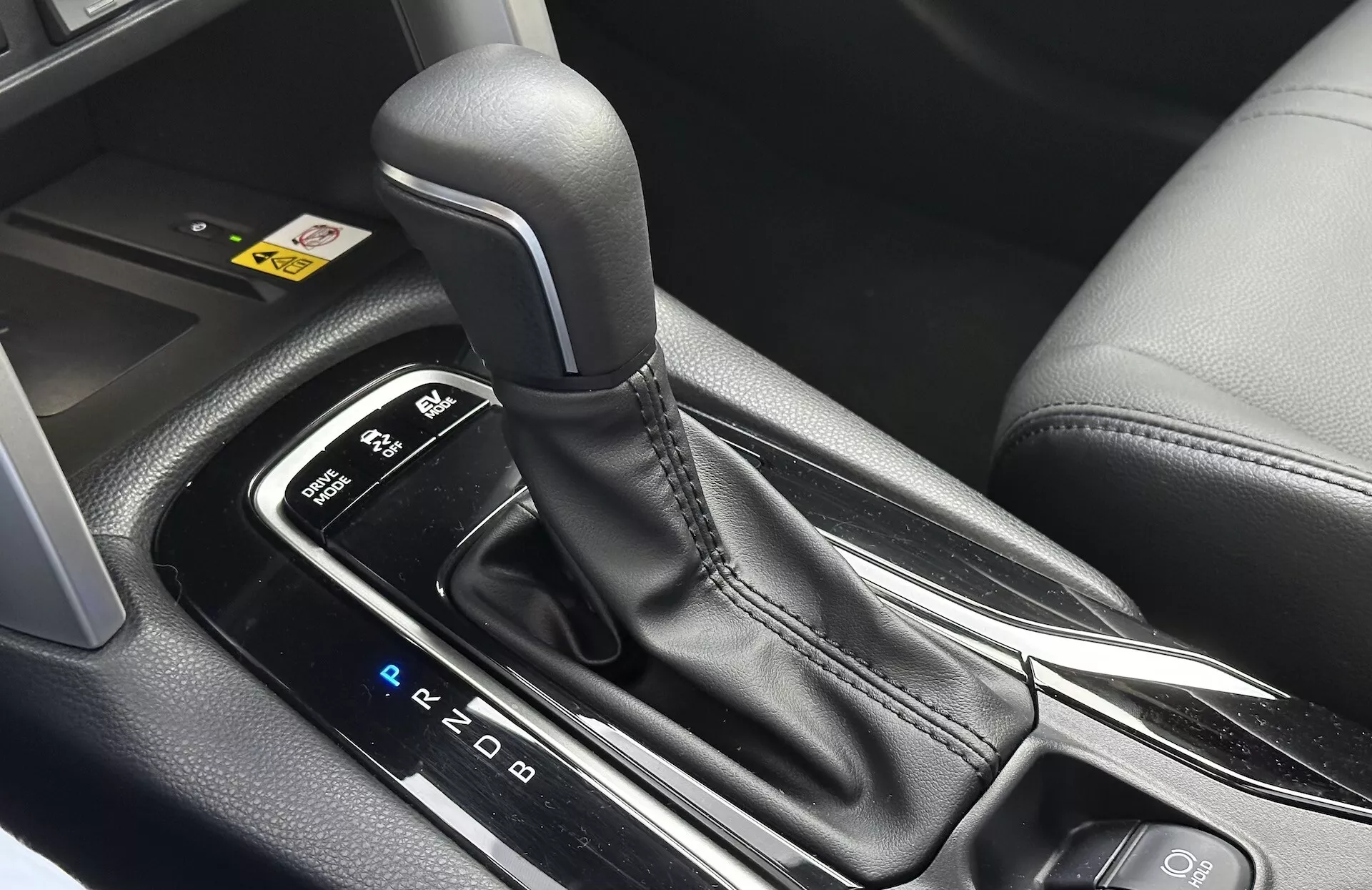 Detailný interiérový pohľad na Toyota Corolla Cross 2022 zobrazujúci automatickú radicu páku s viditeľným rozložením PRND, obklopenú elegantnou stredovou konzolou s čiernymi plastovými a kovovými prvkami, zdôrazňujúci ergonomický dizajn a moderné technologické funkcie
