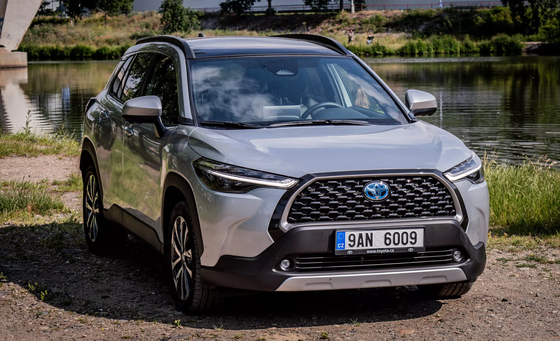 Moderné crossover SUV Toyota Corolla Cross 2022 zaparkované vonku blízko vodnej plochy s jasným pohľadom na jeho prednú mriežku, kapotu, LED svetlomety, predný nárazník a bočný profil vrátane zliatinových kolies a bočných zrkadiel v farbe karosérie