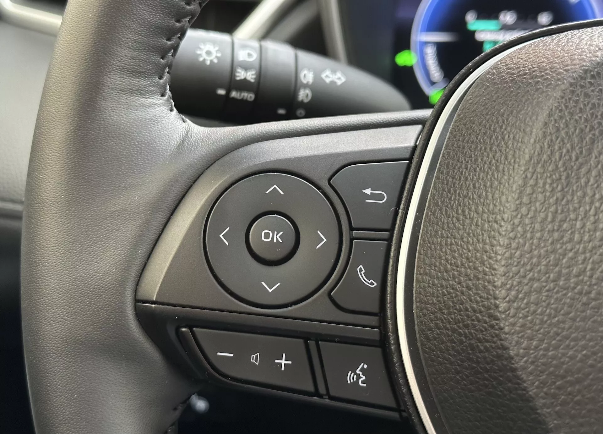 Detailný pohľad na volant Toyota Corolla Cross 2022 zameraný na multifunkčné ovládače na ľavom ramene s textúrou podobnou koži a jasne označenými tlačidlami pre audio, Bluetooth telefónne pripojenie, hlasové príkazy a navigáciu prostredníctvom systému informácií vodiča