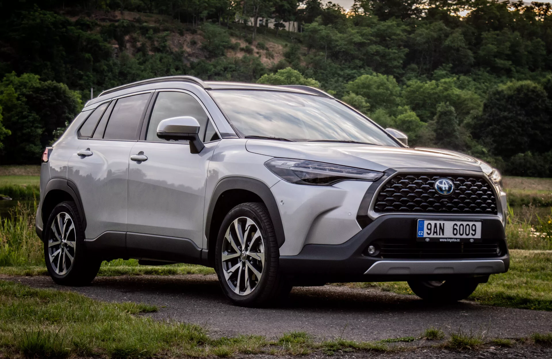 Toyota Corolla Cross 2022 v lesklom kovovom prevedení ukazuje svoj moderný dizajn SUV s detailným predným grilom, emblémom, uhlovými LED svetlometmi, aerodynamickým predným nárazníkom, kontúrovanou kapotou, bočným profilom s litými kolesami s viacerými lúčmi a strešnými nosičmi, snímok zhotovený z nízkeho uhla na zdôraznenie jeho výšky a postoja počas dňa v prírodnom predmestskom prostredí.
