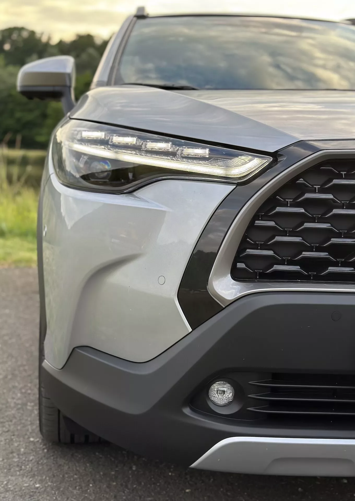Detailný pohľad na prednú ľavú štvrtinu Toyota Corolla Cross 2022 s vyobrazením vzoru šesťuholníkovej mriežky, LED svetlometa, hmlového svetla, predného nárazníka s ochranným krytom, štíhlych karosárskych dielov, bočného zrkadla s integrovaným smerovým signálom a metalického laku