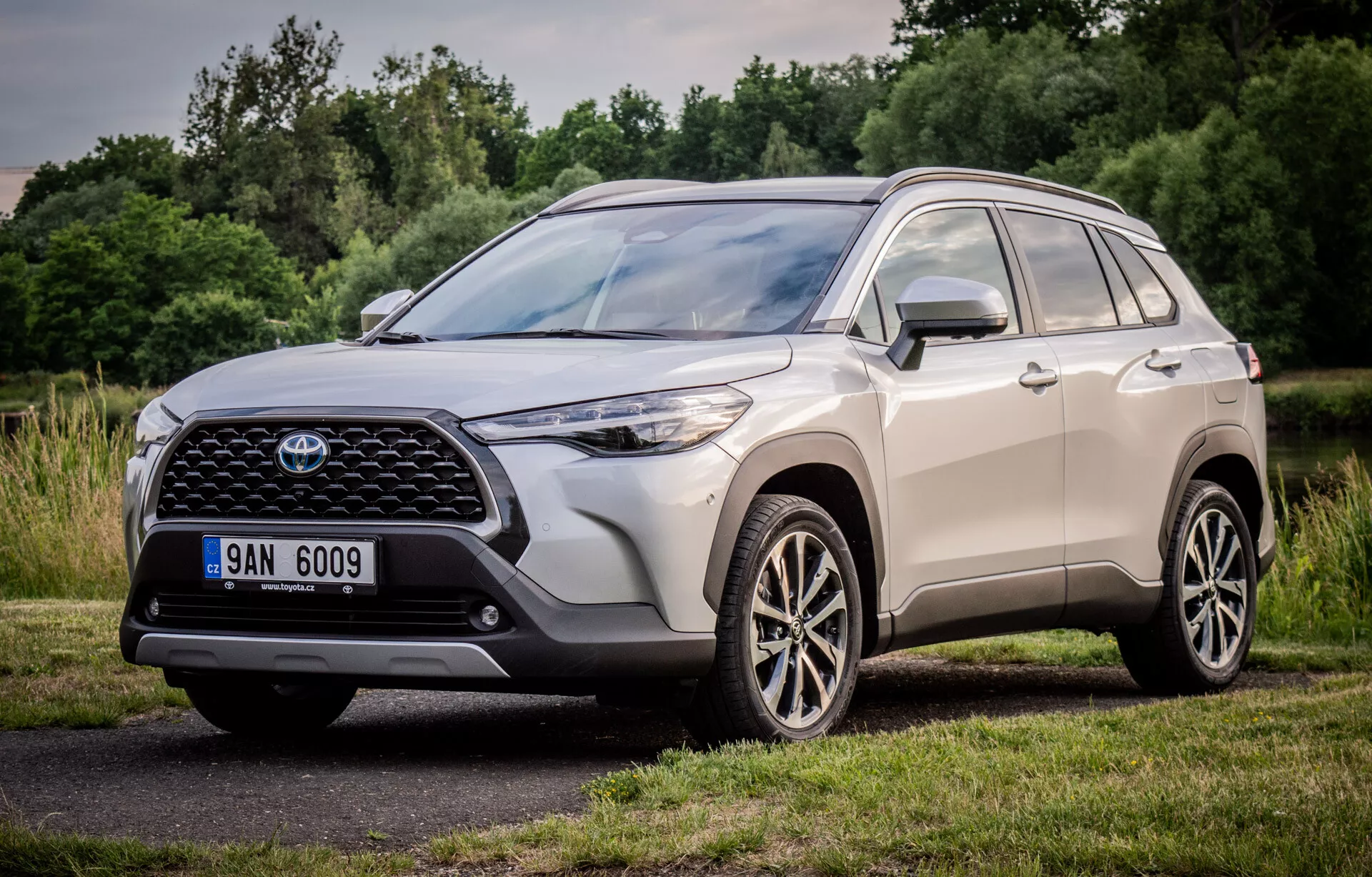 Toyota Corolla Cross 2022 predstavuje svoj dynamický dizajn s trojštvrťovým pohľadom spredu a z boku, zdôrazňuje veľkú vzorovanú prednú mriežku, štíhle LED svetlomety a viaceré zliatinové kolesá, zaparkované v prírodnom parkovom prostredí