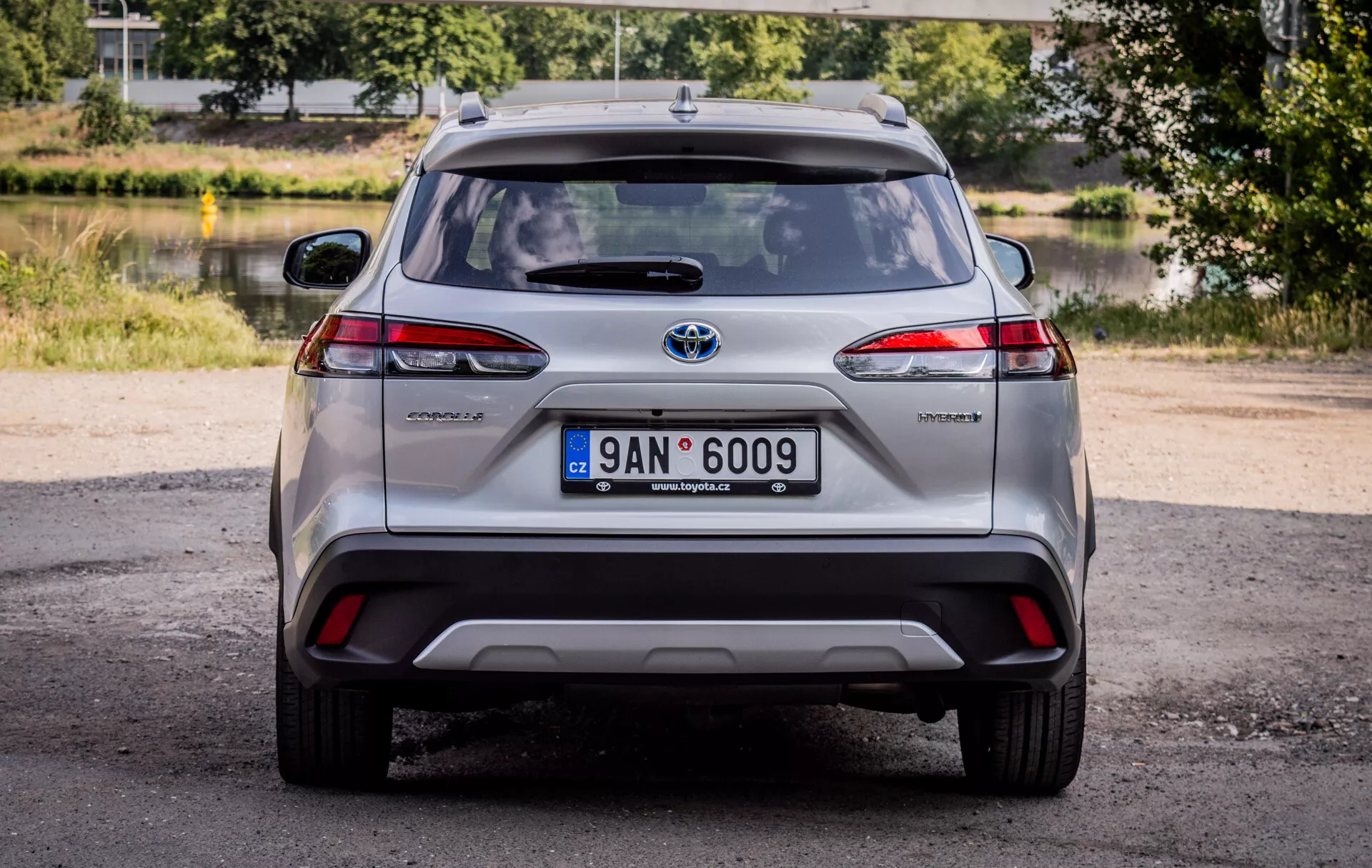 Zadný pohľad na Toyota Corolla Cross 2022, zobrazujúci LED zadné svetlá, odznak hybrid a chrómový emblém Toyota s prírodným denným osvetlením a scenickým pozadím zelene a vody