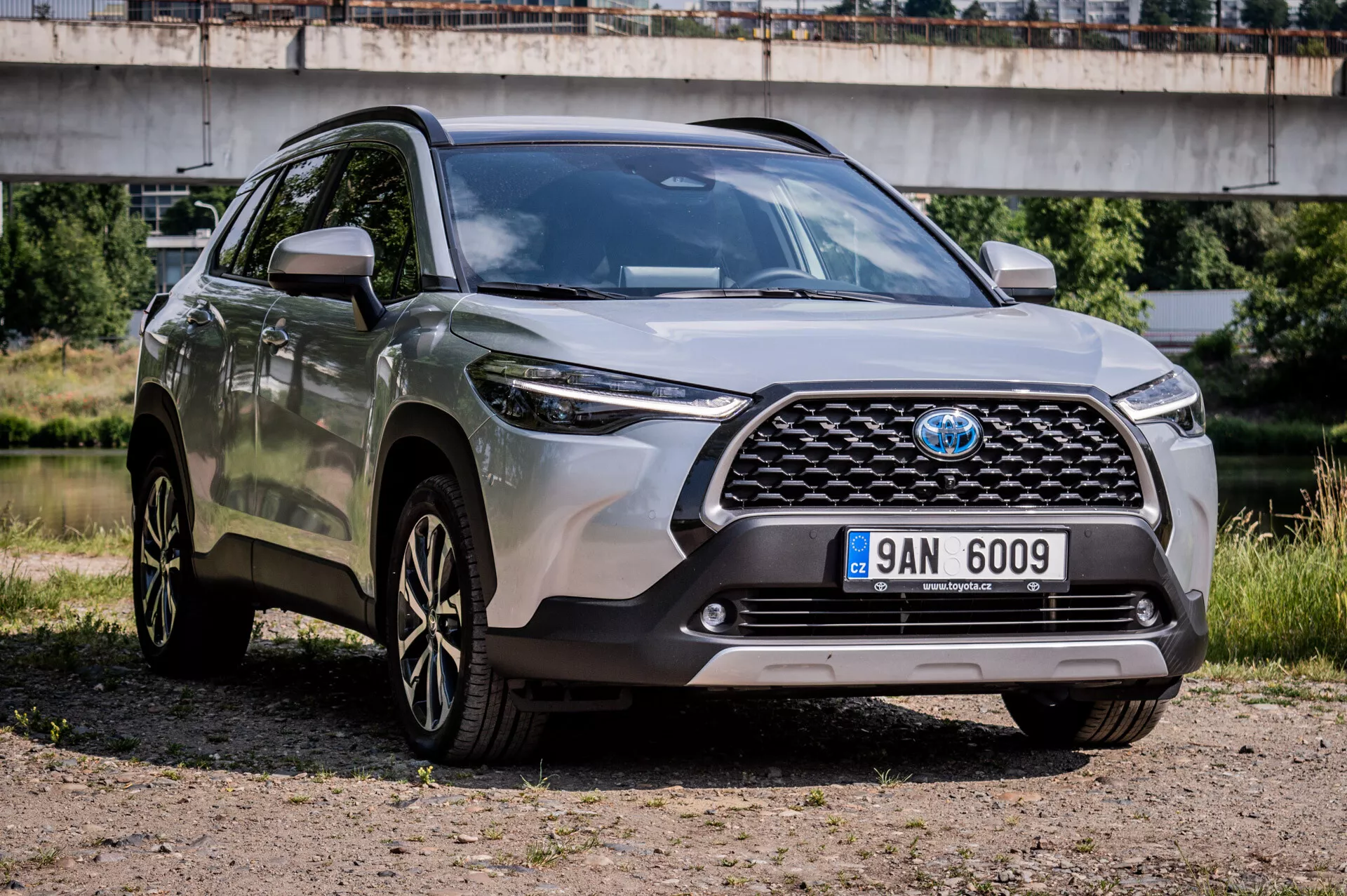 Toyota Corolla Cross 2022 zaparkovaná na štrku ukazuje svoj robustný dizajn, športové disky z ľahkej zliatiny s viacerými paprskami, pokročilé LED svetlomety a vysokú svetlú výšku z pohľadu z troch štvrtín na prednú mriežku, nárazník a prvky na strane vodiča s mostom na polomestskom pozadí