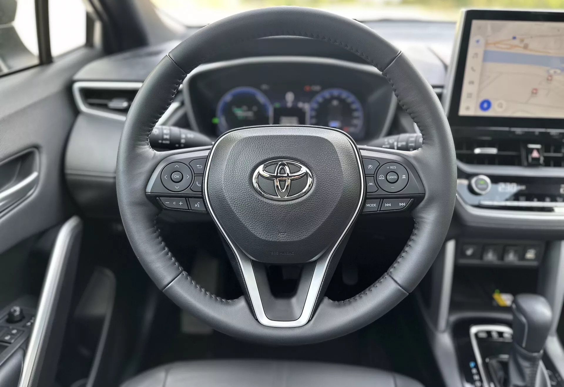 Interiér Toyoty Corolla Cross 2022 so zameraním na volant s logom Toyoty, integrovanými ovládačmi pre audio, hovory a tempomat, za ktorým čiastočne vidieť digitálny prístrojový panel so speedometrom, otáčkomerom a multifunkčným displejom, vrátane väčšieho centrálného displeja na palubnej doske pre GPS navigáciu, všetko na pozadí súčasného dizajnu z tmavých materiálov.