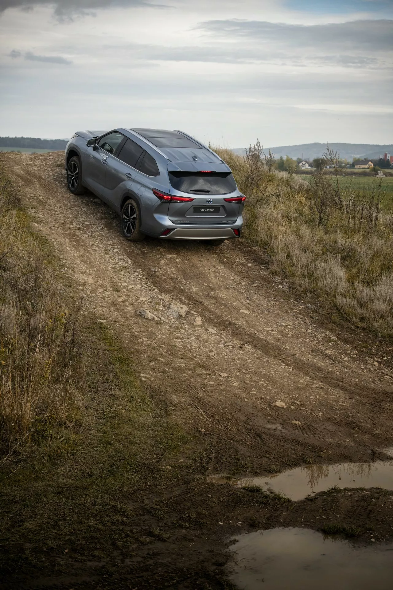 Toyota Highlander 2021 tmavý hatchback zachytený z vysokého uhla na nespevnenej ceste, predstavuje zadný trojštvrťový pohľad, športový dizajn a potenciál pre ľahkú jazdu v teréne uprostred vidieckej krajiny