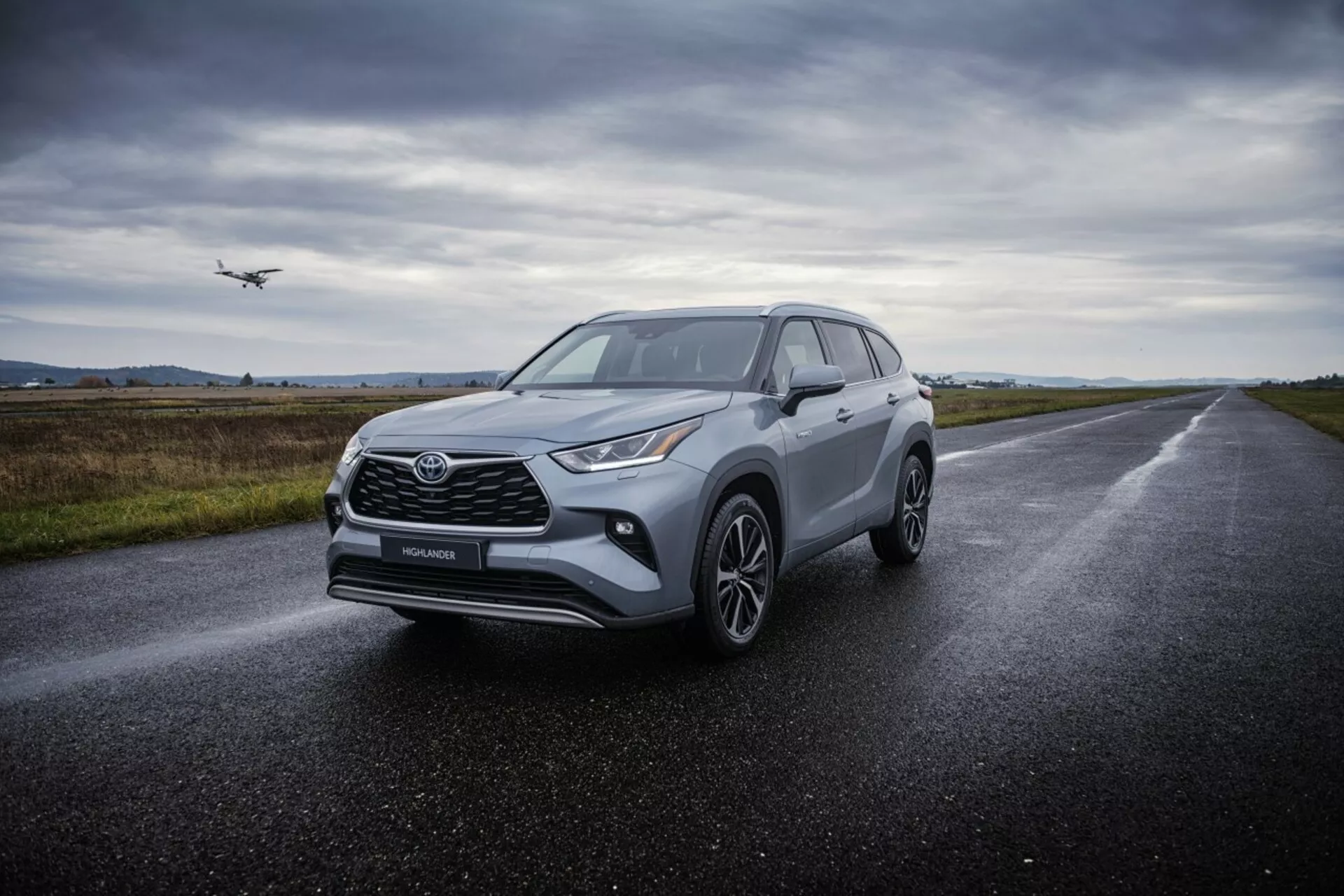 Crossover SUV Toyota Highlander 2021 na mokrej asfaltovej ceste, predvádzajúce svoje svetlomety ostrých uhlov, výraznú prednú mriežku, zliatinové kolesá s viacero lúčmi a aerodynamický dizajn pod rozptýleným zamračeným osvetlením