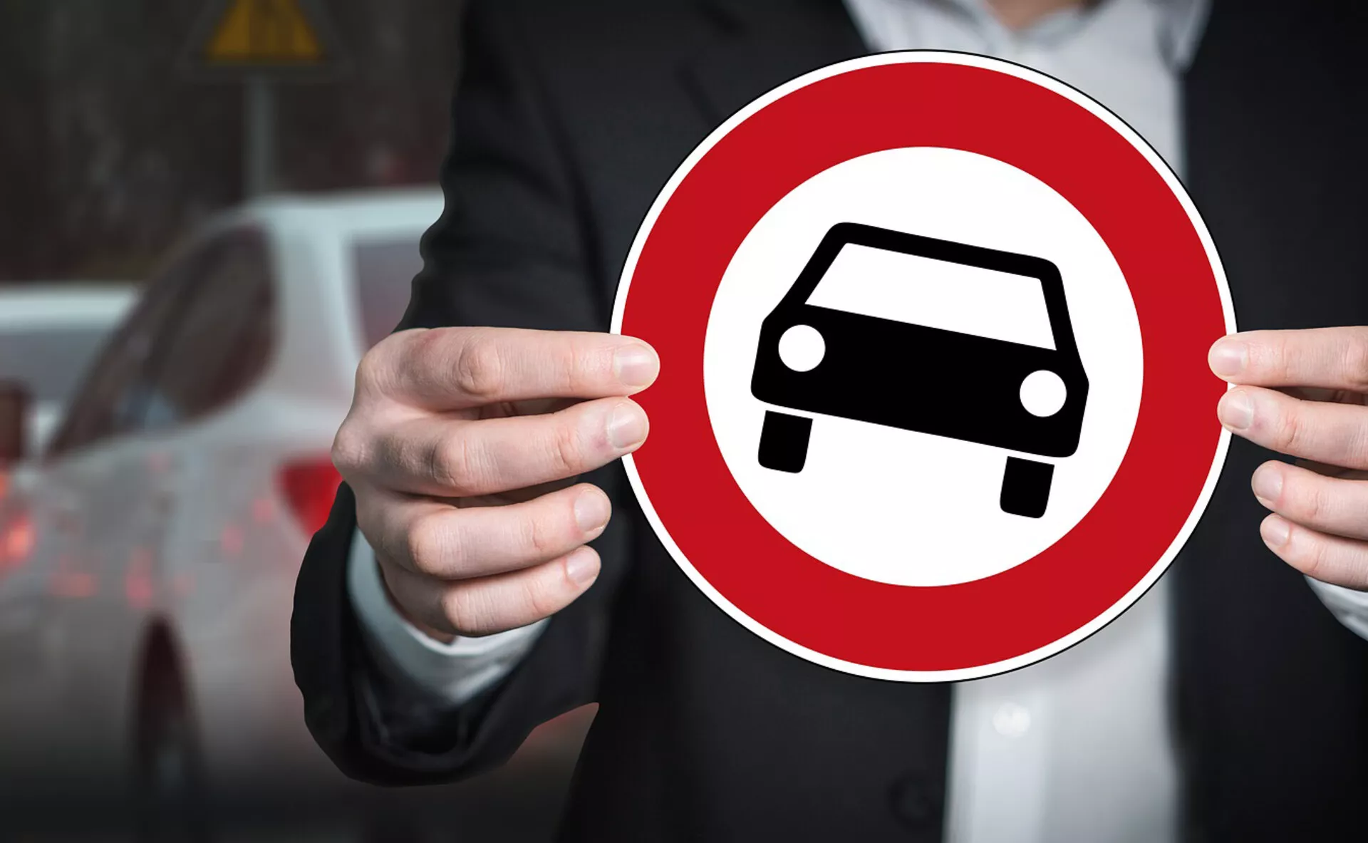 Osoba držiaca červený kruhový značka s bielou siluetou auta, symbolizujúca všeobecné zastúpenie automobilu na rozostrenom mestskom pozadí