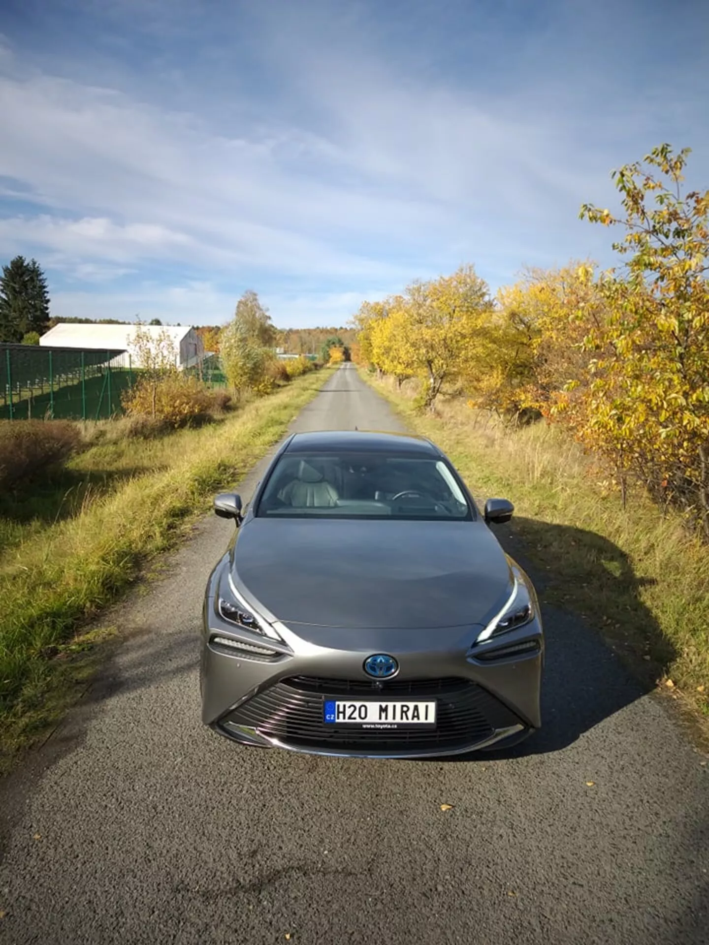 Toyota Mirai 2021 odfotená z prednej strany pod miernym uhlom, zobrazujúca jej štíhle LED svetlomety a výraznú mriežku s centrálnym emblémom, ktorý môže obsahovať senzorovú technológiu, na pozadí polovidieckej krajiny pri prirodzenom dennom svetle, predvádzajúca pokročilú technológiu vodíkových palivových článkov a aerodynamický dizajn