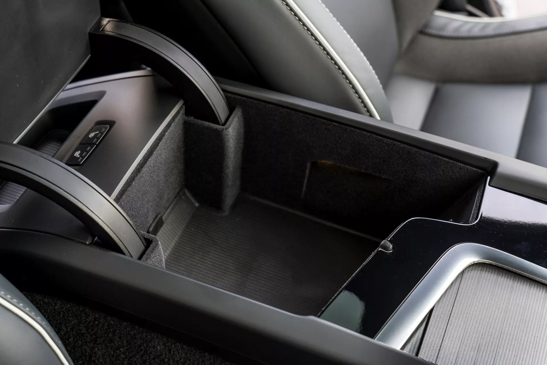 Detailný pohľad na oblasť stredovej konzoly v interiéri modelu Volvo XC60 2021, ktorý ukazuje otvorený odkladací priestor s plstnatým materiálom, dvojúrovňové skladovanie s menším horným podnosom, koženú lakťovú opierku a kovové ozdoby pridávajúce eleganciu. Pri okraji obrázka je viditeľná časť oblasti radiacej páky s koženým poťahom.