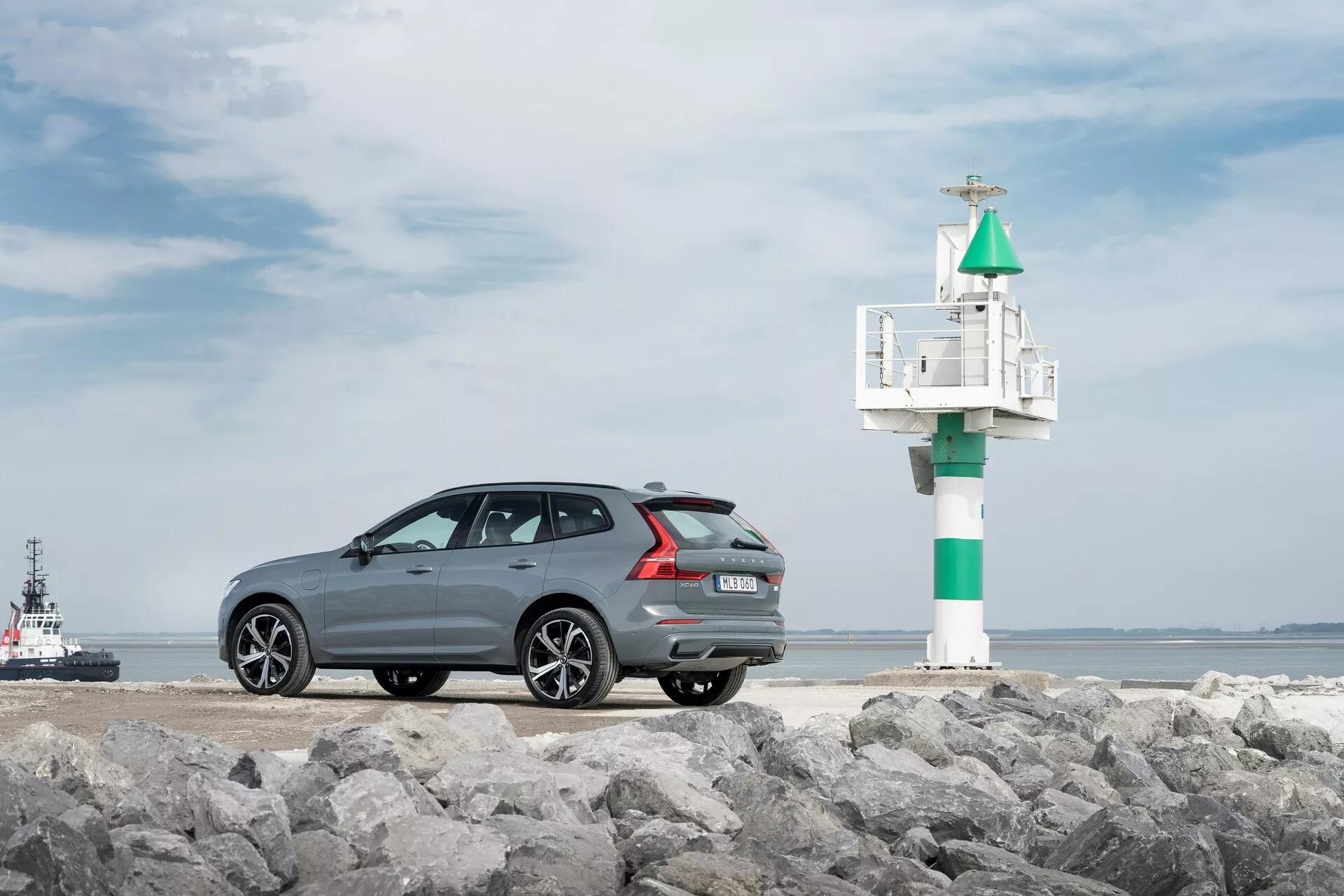 Fotografia modelu Volvo XC60 2021 druhej generácie po facelifte, zachyteného z trojštvrťového zadného uhla na kamenistom povrchu, s námornou scenériou v pozadí. Tmavošedé auto s kovovým lakom má elegantnú, športovú strešnú líniu, viacpaprskové zliatinové kolesá, zadné parkovacie senzory a dvojitý výfuk. Na pravej strane auta je vidieť malý maják alebo navigačný signál, s remorkérom alebo pilotným člnom vo vode, čo zvyšuje pobrežnú atmosféru.