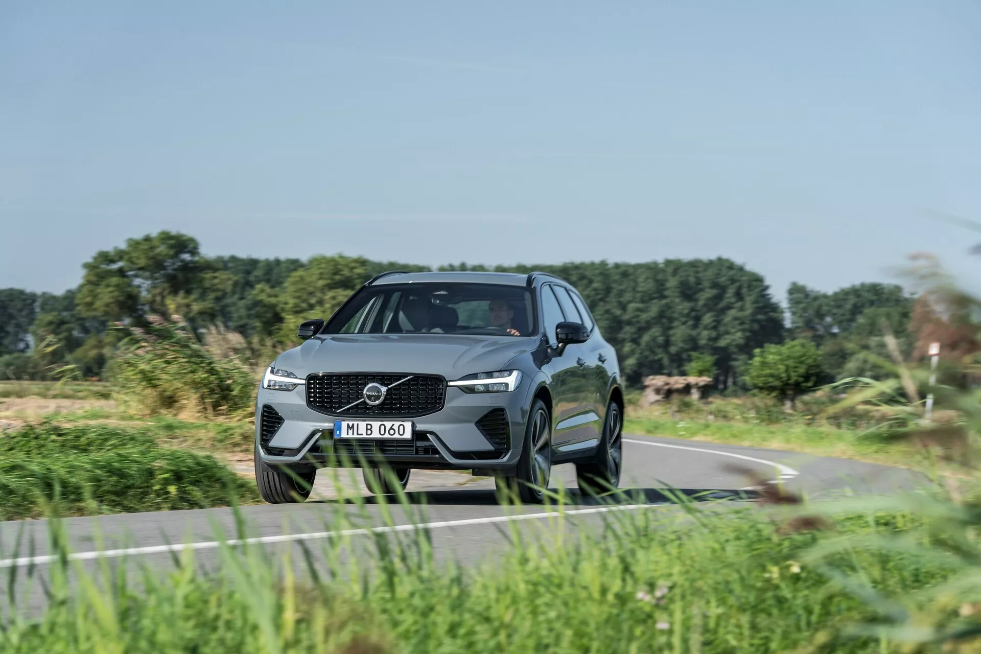 Moderné SUV, Volvo XC60 2021 (facelift 2. generácie), zachytené z nízkeho predného trojštvrťového uhla, zdôrazňujúce dynamický postoj a dizajnové prvky. Výrazný dizajn mriežky s veľkým emblémom, elegantné LED svetlomety a tvarovaný nárazník s integrovanými sacími otvormi. Aerodynamický, luxusný vzhľad s kontúrami, vysokou svetlou výškou, veľkými viacramennými zliatinovými kolesami, nízkoprofilovými pneumatikami, zrkadlami so zabudovanými smerovkami, klesajúcou siluetou strechy a tónovanými oknami. Naznačuje prítomnosť moderných technologických a bezpečnostných prvkov, jazdiaci po vidieckej ceste s rozmazanou vegetáciou, čo naznačuje pohyb.