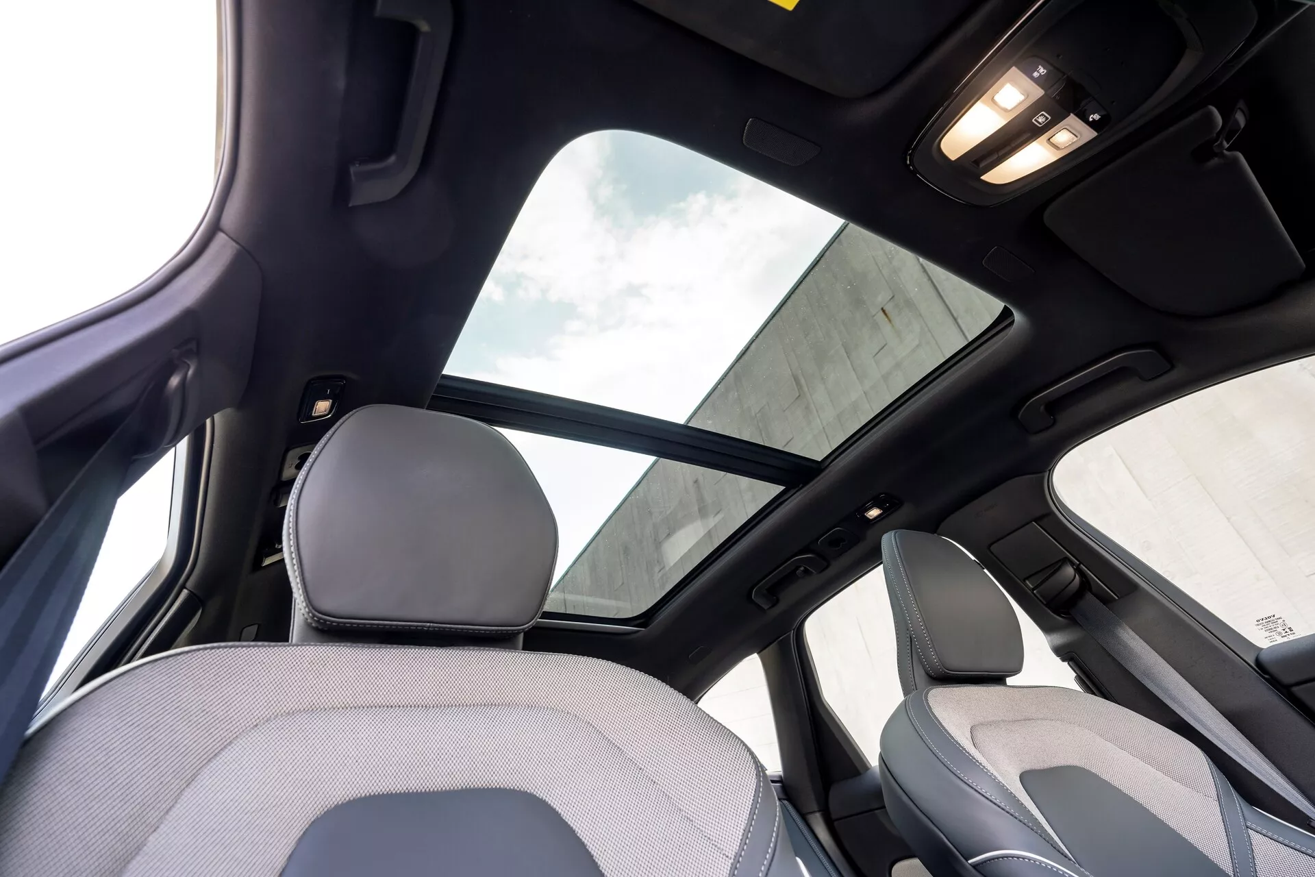 Interiér Volvo XC60 2021 facelift 2. generácie z pohľadu zadného sedadla, zameraný na hornú časť kabíny. Významné prvky zahŕňajú veľké panoramatické strešné okno s tieniacou clonou, čiernu stropnicu indikujúcu vysokú úroveň výbavy alebo luxusný balík a osvetlenie kabíny na strope. Záber zachytáva časť hlavovej opierky a hornej časti operadla sedadla spolujazdca, stĺpik B a rukoväť nad zadnými dverami.