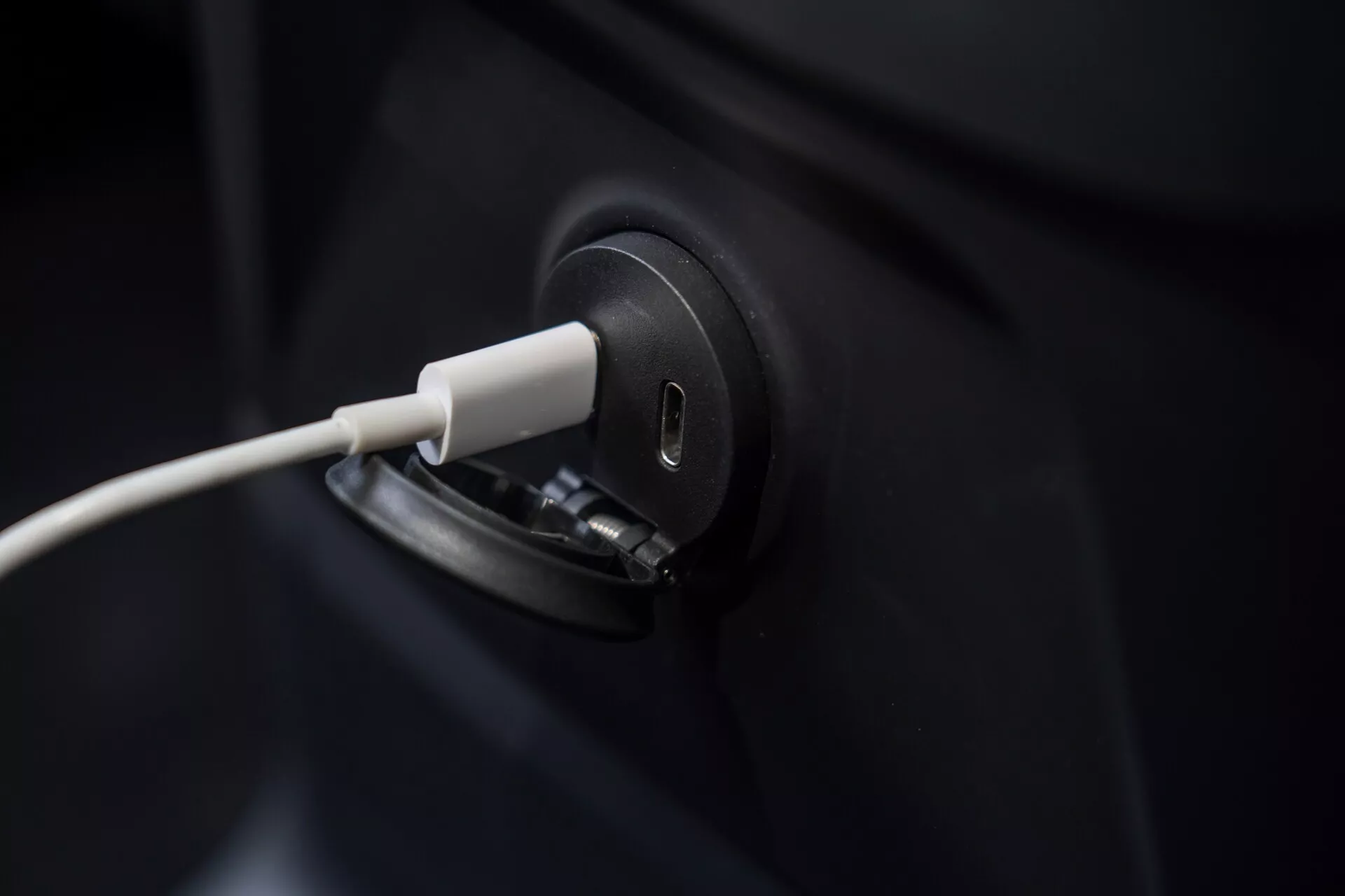 Detailný záber na nabíjací port elektrického vozidla Volvo XC60 2021 s pripojeným káblom, zdôrazňujúci elegantný a moderný dizajn. Záber je úzko zameraný na nabíjací port, pričom väčšina tmavého karosérie vozidla nie je viditeľná. Rozmazané pozadie zvýrazňuje presnosť nabíjacieho procesu EV, symbolizujúce zelené technológie a udržateľnú dopravu.