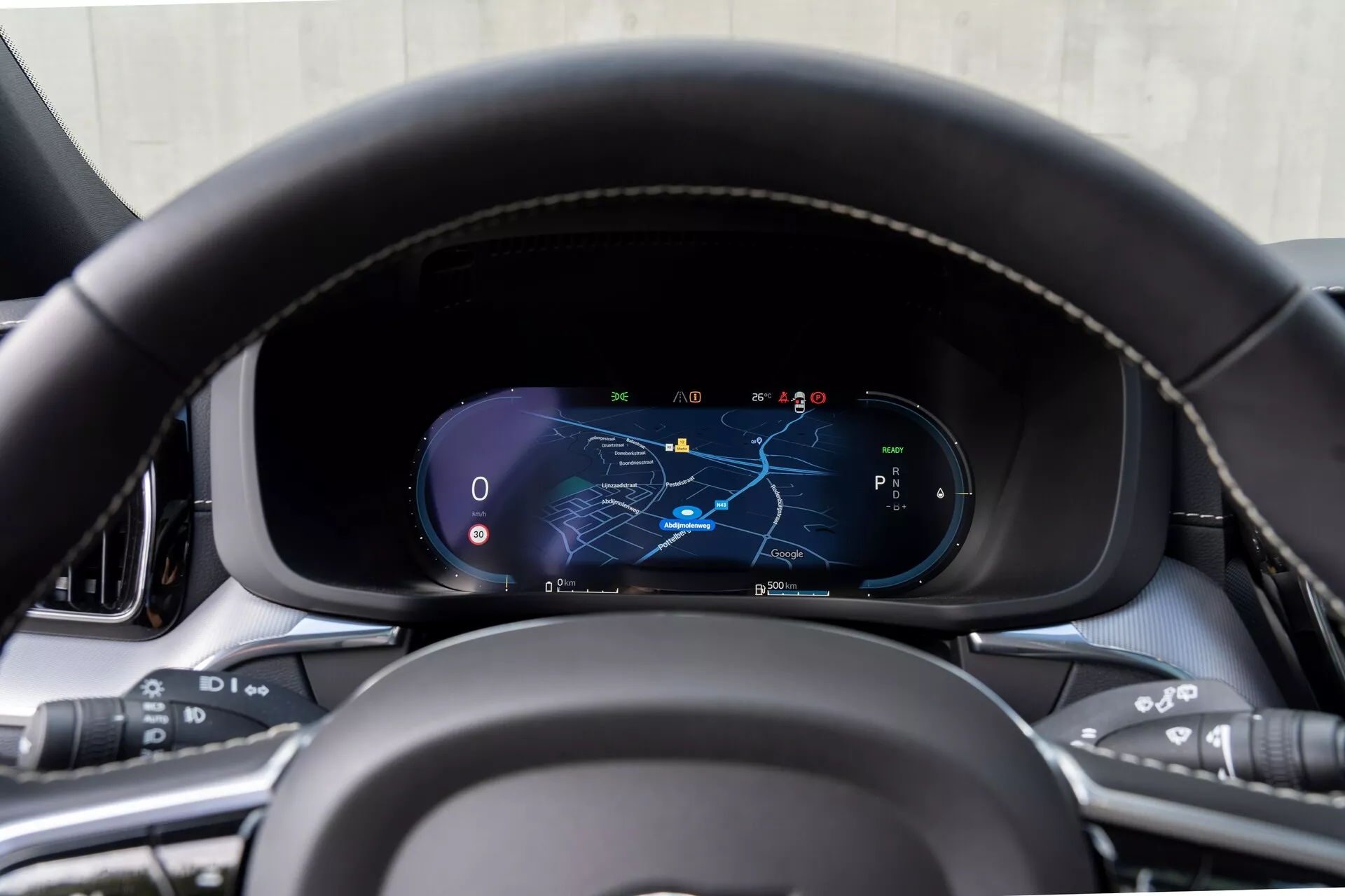 Zblízka pohľad na palubnú dosku Volvo XC60 z roku 2021 (2. generácia-facelift) z pohľadu vodiča, zameraný na digitálny prístrojový panel s koženým volantom s detailmi šitia a multifunkčnými tlačidlami na médiá, hlasové príkazy, tempomat a systémy asistencie vodiča. Digitálny displej zobrazuje mapu s vyznačenou navigačnou trasou, tachometer, otáčkomer a výber prevodového stupňa v parkovacom režime, obklopený prémiovými materiálmi interiéru.