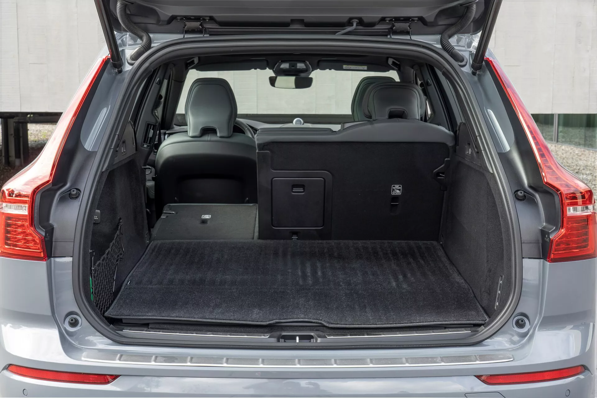 Obraz zobrazujúci zadný nákladový priestor vozidla Volvo XC60 z roku 2021 (2. generácia facelift), fotografovaný z priameho uhla s otvoreným kufrom. Stredne vzdialený záber zachytáva celú šírku nákladového priestoru, ktorý je pokrytý čiernym kobercovým materiálom. Na ľavej strane je malý úložný priestor so sieťkou. Pravé zadné sedadlo je sklopené, odhaľujúc delené sklopné sedadlá, zatiaľ čo ľavé sedadlo zostáva vo zvislej polohe s opierkou hlavy. Nad zadnými sedadlami je umiestnený zasúvací kryt nákladu, ktorý predstavuje skladovaciu všestrannosť vozidla.