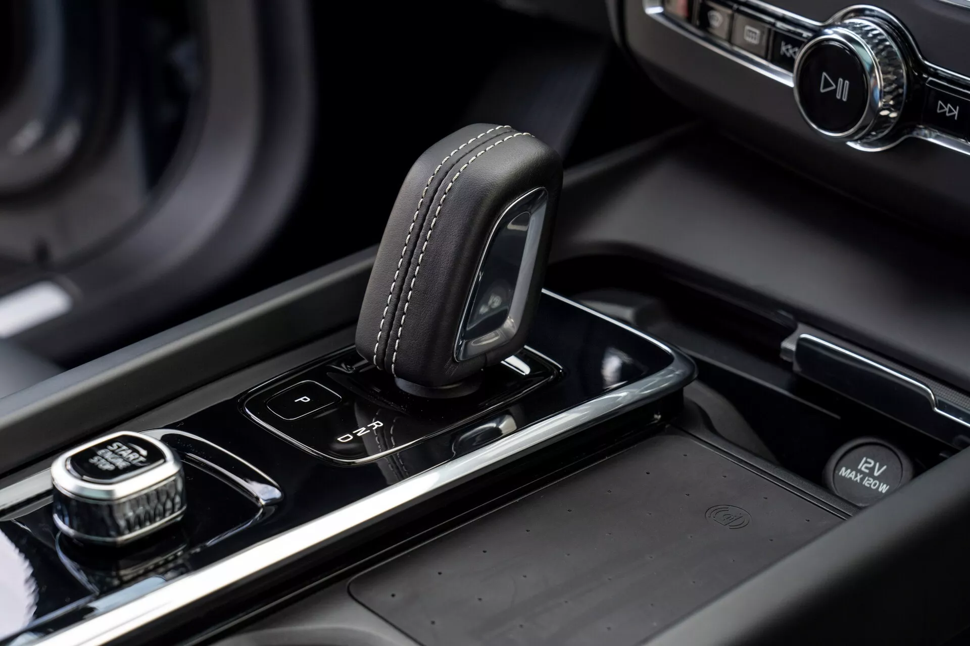 Detailný záber na stredovú konzolu vozidla Volvo XC60 z roku 2021 (2. generácia, facelift). Obrázok zvýrazňuje volič prevodovky s koženým povrchom a detailným šitím, umiestnený na lesklom čiernom povrchu. Napravo sa nachádza ciferník s vrúbkovaným okrajom, pravdepodobne pre navigáciu v infotainmente, s tlačidlom pre voľbu stlačením. Vedľa voliča prevodovky sú tlačidlá pre parkovacie senzory a automatický start/stop systém. Stredová konzola obsahuje kovové prvky a v rozmazanom pozadí sa nachádza osvetlený infotainment displej, čo zdôrazňuje moderný, vysoko kvalitný dizajn a spracovanie vozidla.