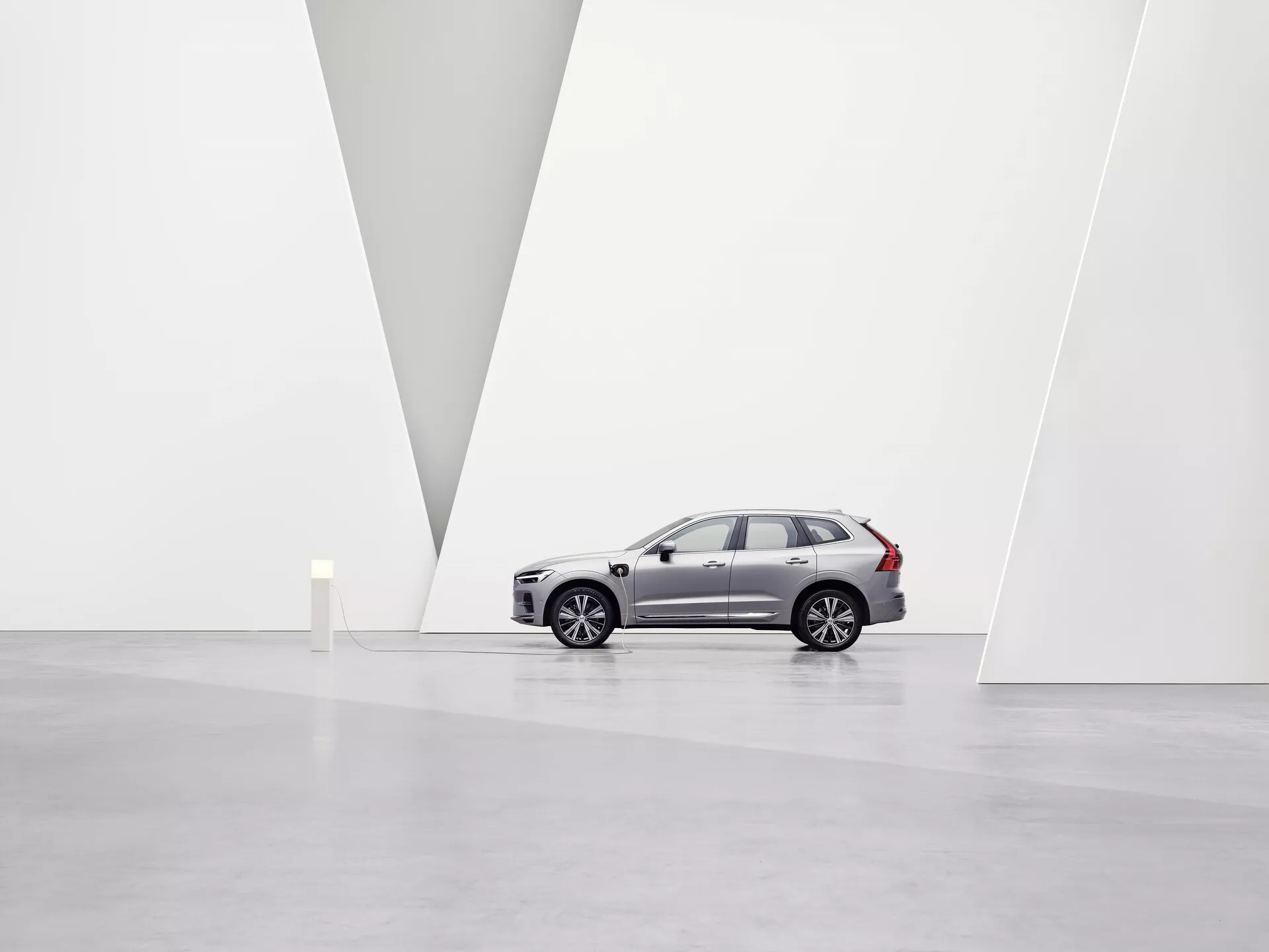 Na obrázku je Volvo XC60 2021 druhej generácie facelift SUV umiestnené v priestrannej, minimalistickej umeleckej galérii s bielymi stenami a lesklou podlahou. Vozidlo zobrazené z profilu smerom doľava zvýrazňuje hliníkové kolesá, dizajnové línie bočných dverí, bočné zrkadlá, zatemnené sklá a charakteristické strešné lišty. Z mierne nízkeho uhla sú viditeľné predná mriežka, LED svetlomety, integrované smerové svetlá a parkovacie senzory na prednom nárazníku. Čisté prostredie a V tvarová delička na stene zdôrazňujú dizajn auta a jeho prémiovú pozíciu na trhu pod mäkkým, rozptýleným osvetlením.