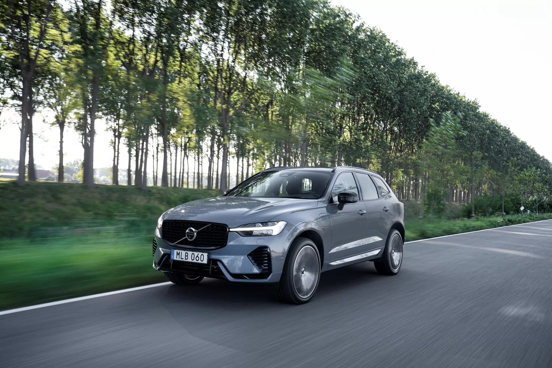 Obrázok zobrazuje pohybujúci sa Volvo XC60 2021 (facelift 2. generácie) zachytený z predného trojštvrťového uhla, ktorý ukazuje vodičovu stranu auta. Tento moderný luxusný SUV má výrazný elegantný dizajn a emblém na prednej mriežke. Záber z mierne zostupného uhla zahŕňa kapotu, nárazník, mriežku, svetlomety a kolesá. Vozidlo tmavej metalízy, pravdepodobne sivej alebo čiernej, s lesklým povrchom, disponuje hliníkovými kolesami, LED svetlami a aerodynamickou karosériou. Rozmazané pozadie ulice so stromami naznačuje pohyb a zdôrazňuje výkonnostné schopnosti vozidla.