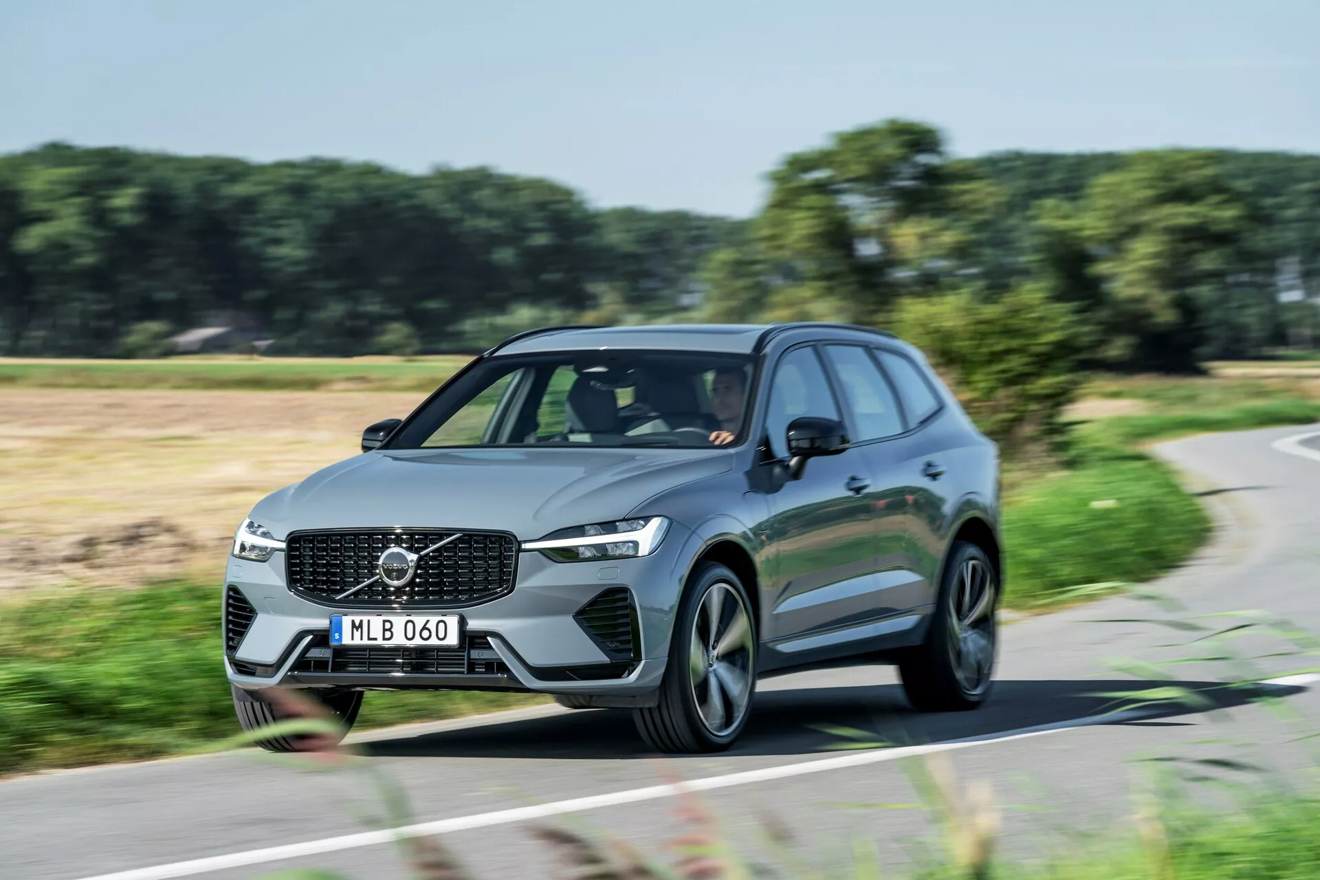 Dynamický denný záber pohybujúceho sa vozidla Volvo XC60 2021 (druhá generácia-facelift) SUV zachytený z trojštvrťového predného uhla. Snímka ukazuje prednú mriežku s výrazným emblémom Volvo, hranaté LED svetlomety, aerodynamické línie a moderné viac-ramenné zliatinové kolesá. Denné svietenie je zapnuté, čo naznačuje, že auto je v pohybe. Fotografia zvýrazňuje elegantný dizajn a cestnú prítomnosť tohto moderného rodinného alebo luxusného úžitkového vozidla.