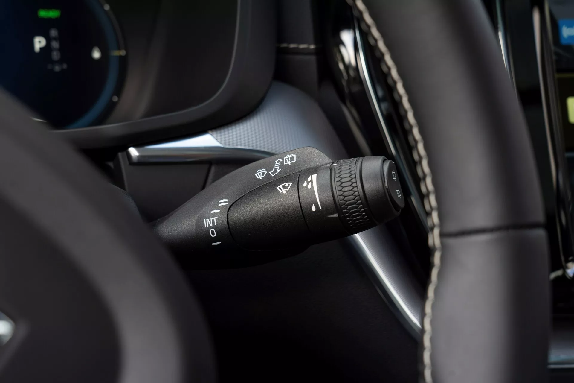 Detailný záber na ovládaciu páčku stieračov Volvo XC60 2021 (facelift druhej generácie) z pohľadu vodiča. Zamerané na rôzne symboly nastavení stieračov: automatická detekcia dažďa, prerušovaná, nízka rýchlosť a vysoká rýchlosť. Na špičke páčky je otočný prsteň na nastavenie intervalu prerušovaných stieračov. Moderný interiér s kombináciou materiálu podobného koži a plastových komponentov s rozmazaným digitálnym displejom v pozadí.