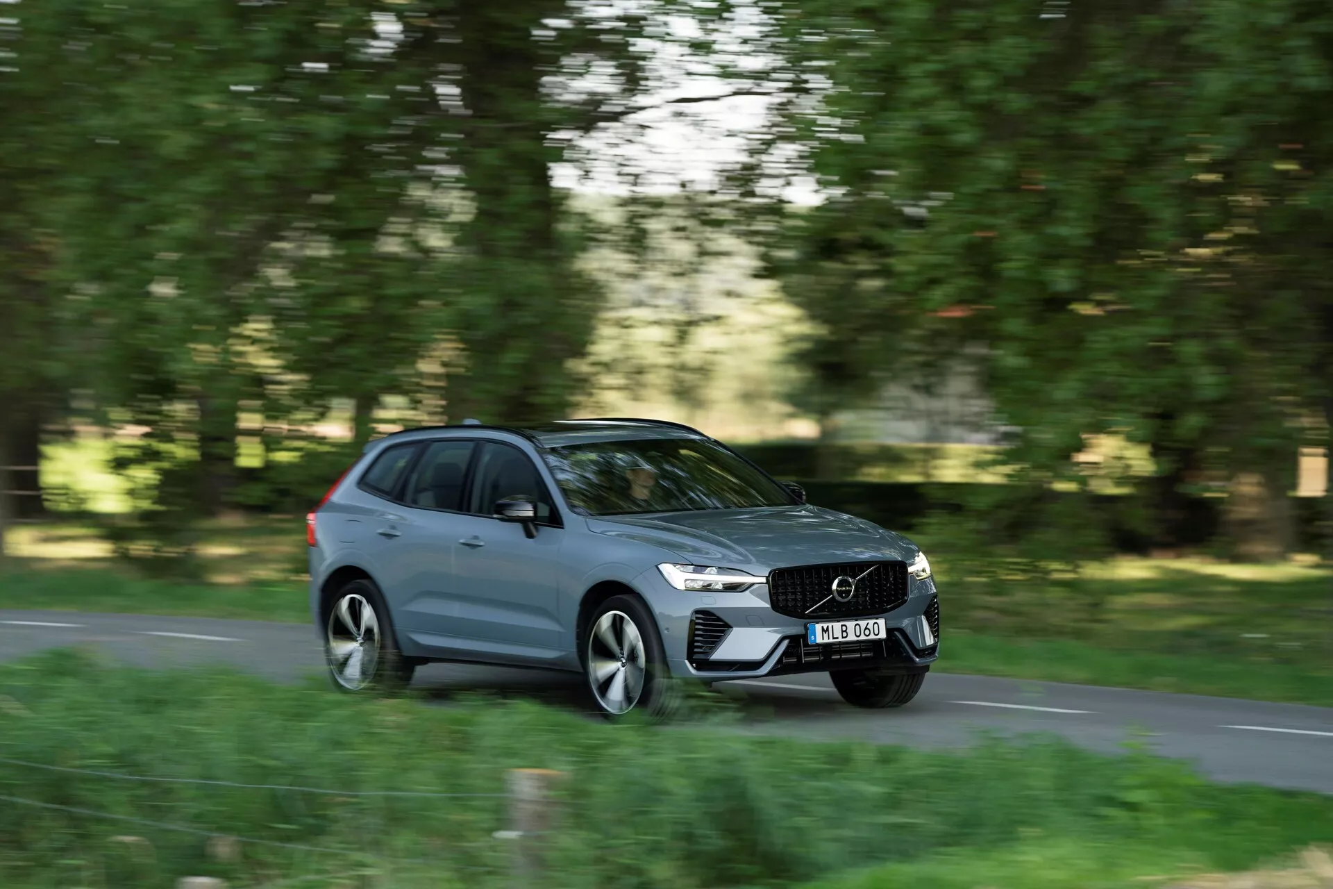 Moderný šedý kombi Volvo XC60 2021 v pohybe, zachytený z bočného uhla na prímestskej alebo vidieckej ceste s zeleňami na obidvoch stranách. Viditeľný predný pravý štvrťpanel, bočná strana a časť zadnej časti. Dynamický pohyb zdôraznený technikou panningu s rozmazaným pozadím a kolesami, ostré detaily vozidla vrátane charakteristickej prednej masky, elegantných svetlometov, aerodynamických línií a zapustených kľučiek dverí. Dizajn viditeľný zo strednej vzdialenosti, rozmazaná ŠPZ, ukazujúci dizajn a pohyb Volva v prírodnom prostredí.